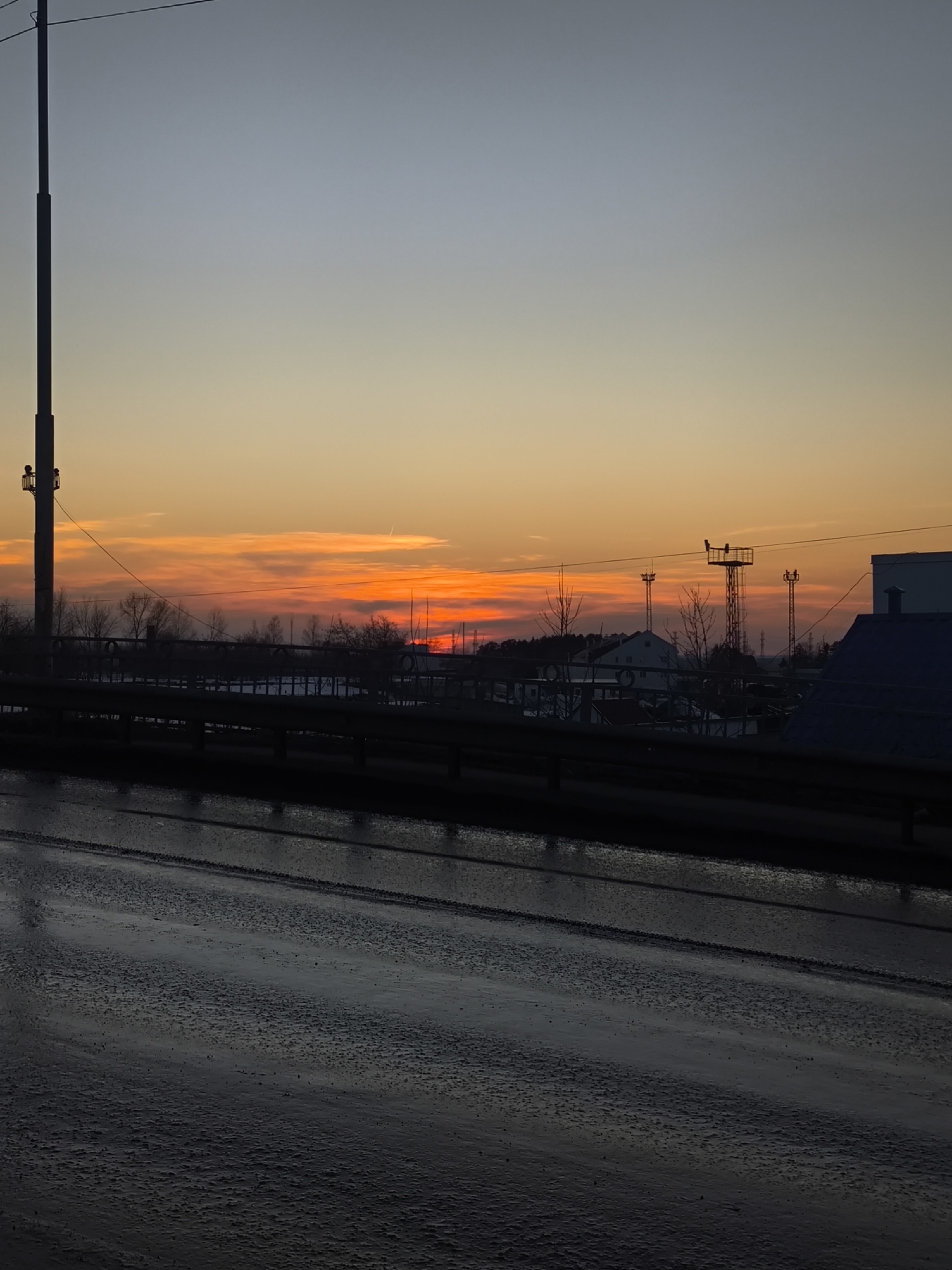 The image captures an urban-industrial landscape at either sunrise or sunset in Chaikovsky, Russia. The foreground consists of a dark, wet asphalt road surface, reflecting ambient light, and showing faint linear markings. A dark, metal barrier or railing extends horizontally across the midground. Behind this barrier, a row of bare trees stands, suggesting a cold season. The background reveals a mix of structures and natural elements: a white building with a dark roof is partially visible among the trees, and several tall, dark metal towers, possibly for communication or power transmission, are silhouetted against the horizon. On the far left, a tall, dark utility pole extends vertically upwards from the lower frame. On the far right, a large white building with a dark, sloped roof is present. The sky above transitions from a light blue-grey at the top to vibrant orange and red tones near the horizon, indicative of a low sun angle. Dark, elongated cloud formations are visible within the orange sky, and a thin contrail is present in the upper-middle sky. There are no individuals or discernible actions occurring within the scene.