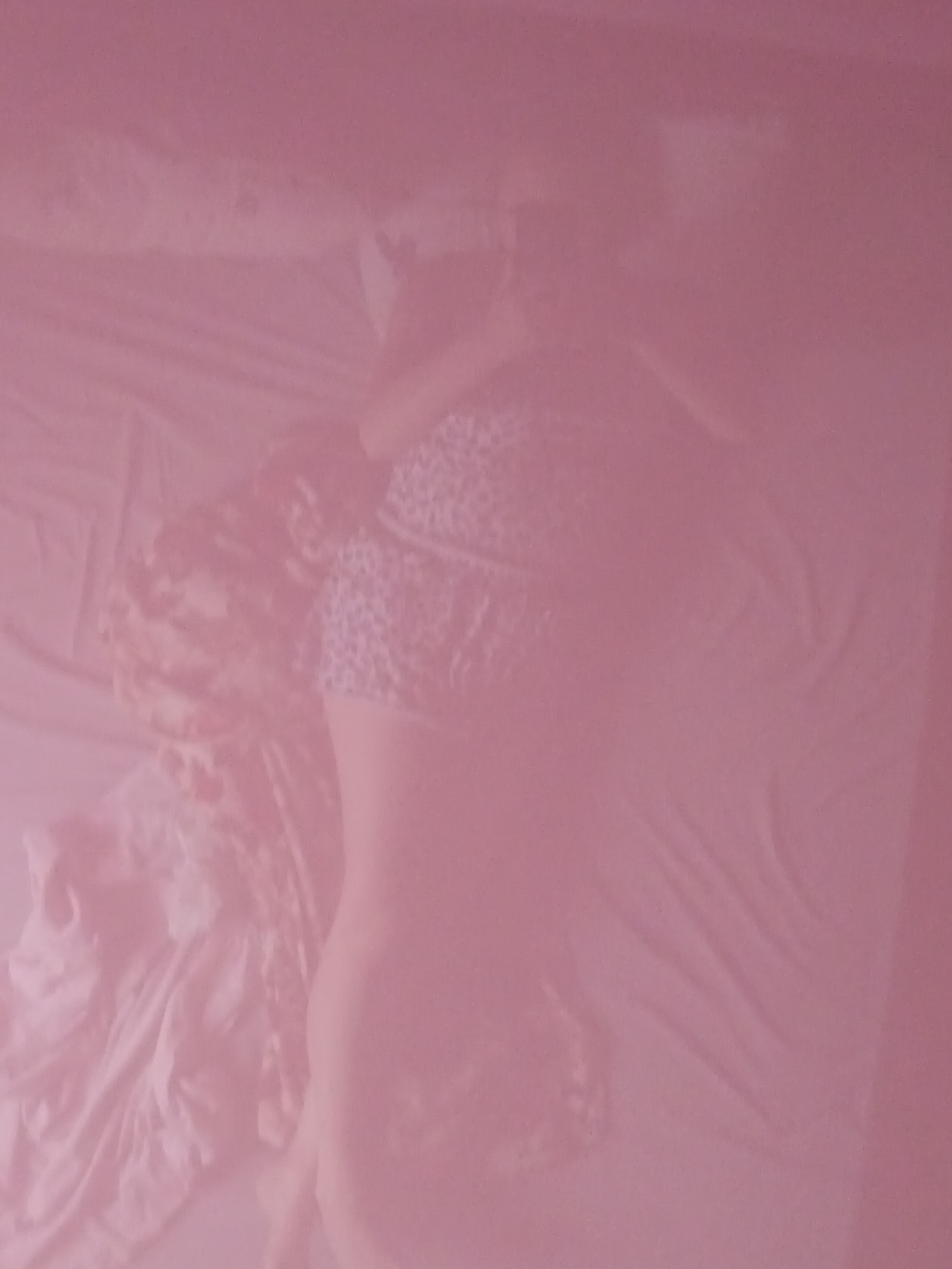 The image depicts a person lying on a bed. The lighting is dim and has a pinkish hue, obscuring finer details. The person is positioned on their side, facing away from the viewer. They appear to be wearing a patterned garment on their upper body. The bed itself is covered with sheets and what seems to be a blanket or throw. The overall impression is that of a private indoor setting. There is no information in the image that could indicate a specific location in the city of Sorochinsk, Russia.