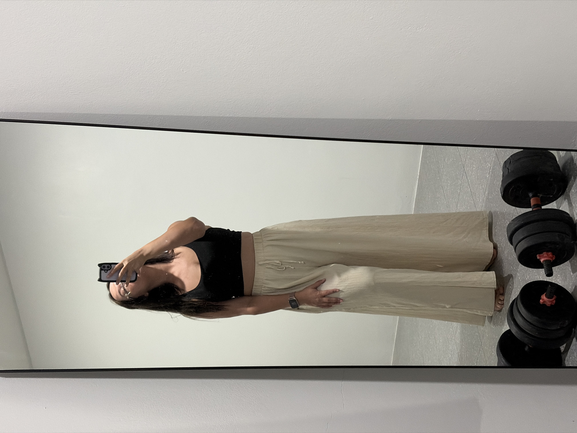 A full-length mirror reflects a young woman standing in what appears to be a gym or workout space. She is wearing a black crop top and wide-legged, cream-colored linen pants with a drawstring. She has a watch on her left wrist and is holding a phone in her right hand, taking a selfie. In the foreground, reflected in the mirror, are two weight sets. The background is a plain white wall. The floor is tiled in a light gray pattern. The location is identified as Baan Chaweng, Thailand.