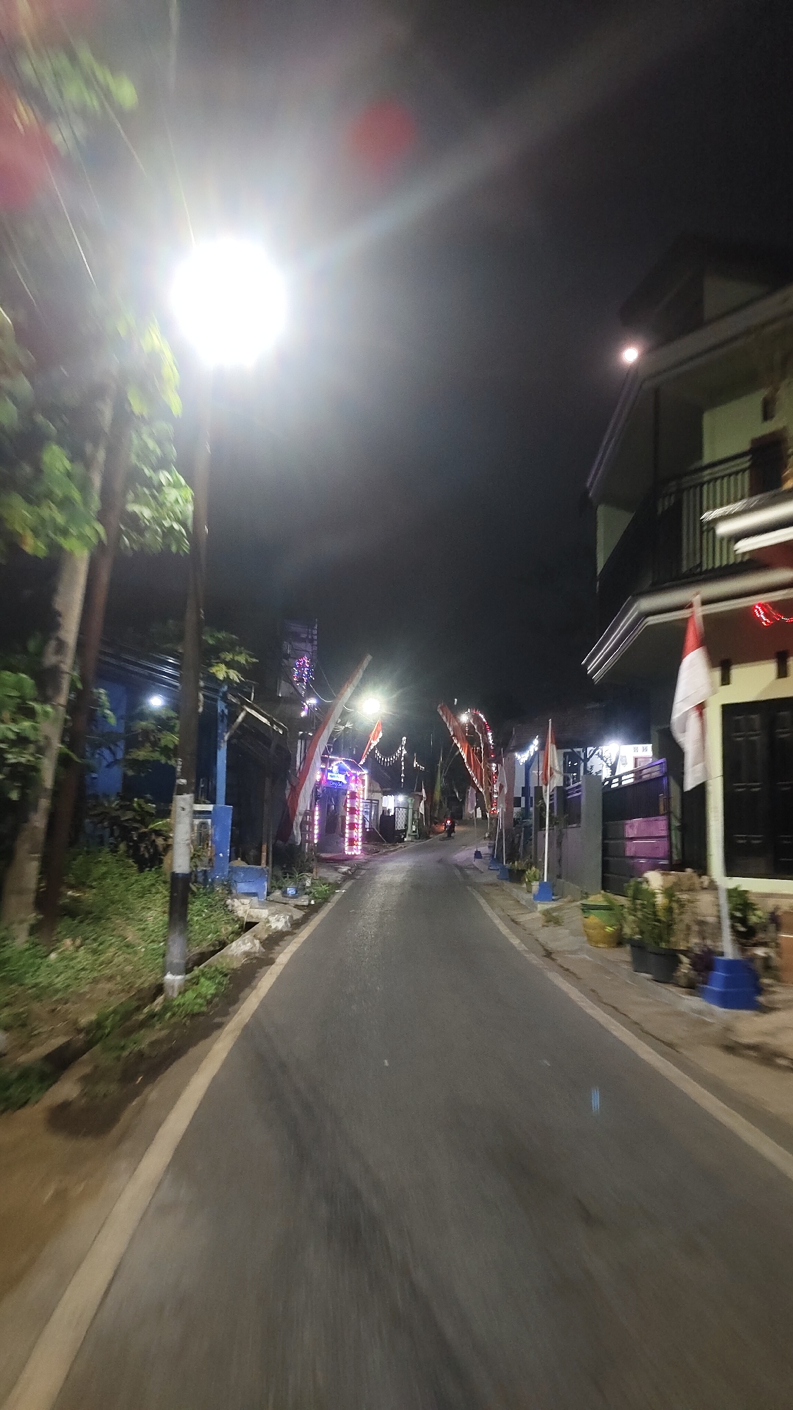 The image depicts a narrow, paved road at night in a residential area, likely in the city of Malang, Indonesia, given the prominent display of Indonesian flags. The road is flanked by buildings and vegetation. A bright street lamp illuminates the foreground, creating lens flare and overexposure. Red and white Indonesian flags are mounted on poles along the sides of the road, some with decorative lights. Buildings with balconies and windows are visible, some adorned with string lights. Potted plants are placed along the walkways. The scene suggests a festive or celebratory atmosphere, possibly related to Indonesian Independence Day. The perspective is from a moving vehicle, indicated by the motion blur.