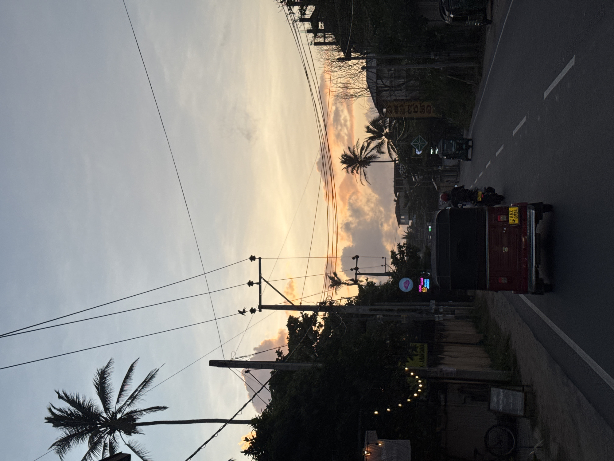 A twilight scene captures a street in Ahangama, Sri Lanka, with a road running through it. The sky displays a gradient of colors, from pale blue at the top to orange and pink hues closer to the horizon, indicating either sunrise or sunset. Several utility poles with a network of overhead wires traverse the frame, some casting shadows on the sky and the surrounding environment.
On the left side of the road, a sidewalk is lined with trees, one of which is a tall palm tree. Decorative string lights with warm yellow bulbs are strung across the scene, illuminating some of the foliage and a sign. A bicycle is parked on the sidewalk, and a sign with an unknown inscription is propped against a wall.
A red auto-rickshaw is the primary vehicle in the foreground, facing away from the viewer and moving down the road. Behind it, a person on a motorcycle is also traveling in the same direction. Further down the road, another auto-rickshaw is visible, as are parts of buildings and more palm trees. The overall impression is a typical street scene in a tropical coastal town during the transition from day to night.