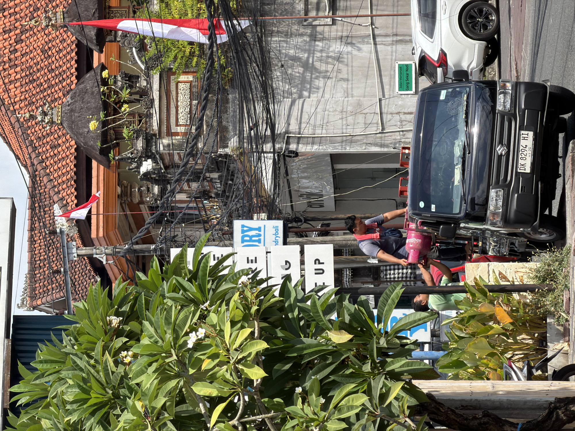 A black Suzuki carry truck is parked on a street, with two men standing near its rear. One man, wearing a blue shirt and red bandana, is standing on the truck and handing a pink gas cylinder to another man in a green shirt who is standing on the ground. The truck is carrying several other gas cylinders, and one is being exchanged. In the background, there are buildings with red tile roofs and intricate stonework, adorned with red and white Indonesian flags. A lush green tree with white flowers is in the foreground, partially obscuring the view of the buildings. Other vehicles, including a white SUV, are visible on the street, and there is a sign in Indonesian that reads "Jalan Mertanadi Gang Sekar Nadi." The scene suggests a commercial street in Indonesia, likely in a tropical climate given the vegetation.