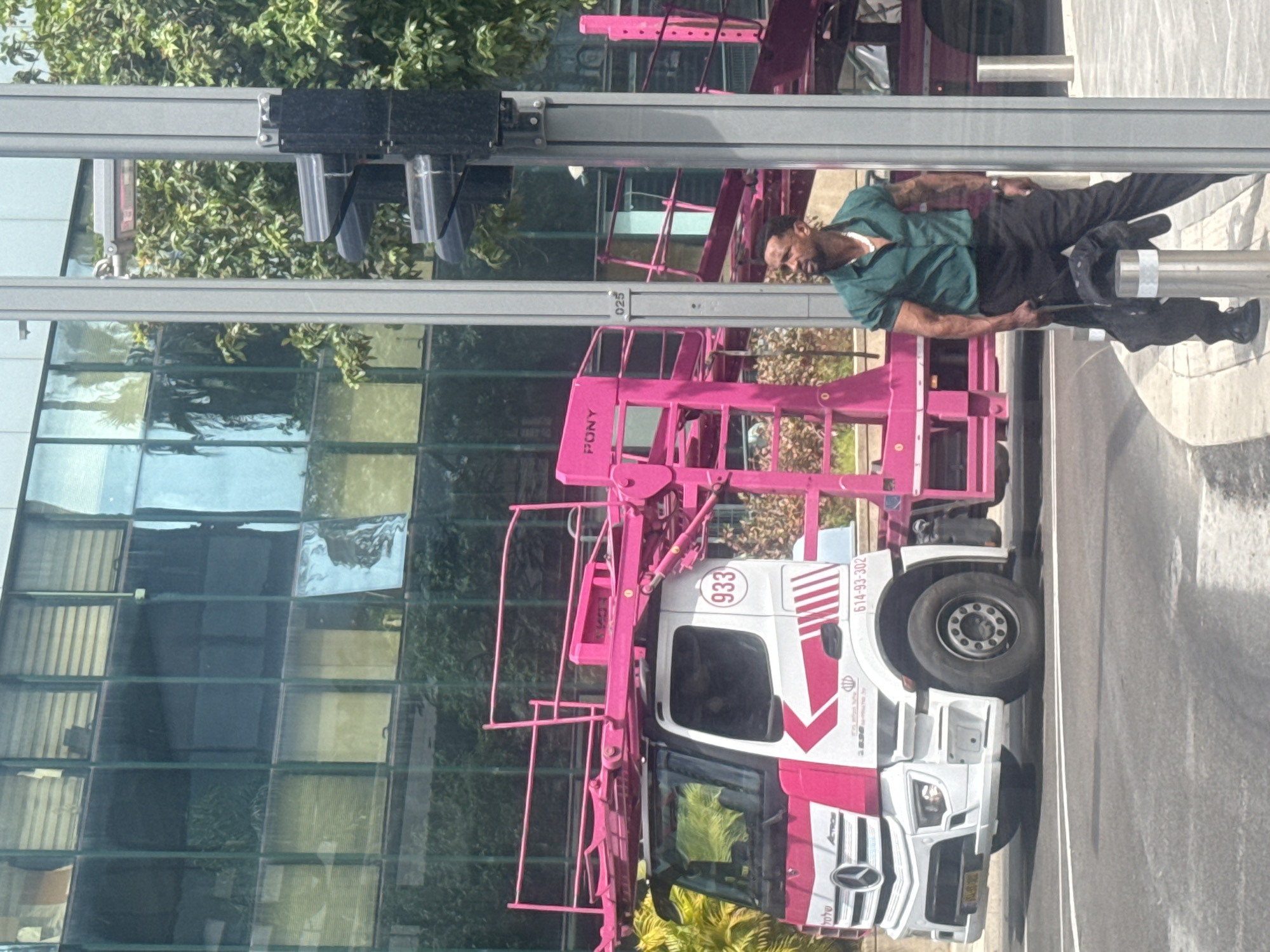 A man in a green shirt and black pants stands on the platform of a pink "PONY" cherry picker parked on a street in Tel Aviv, Israel. The truck is white with pink accents and displays the phone number 614-93-302. The background includes a glass building reflecting trees and the sky, and a street lamp.
