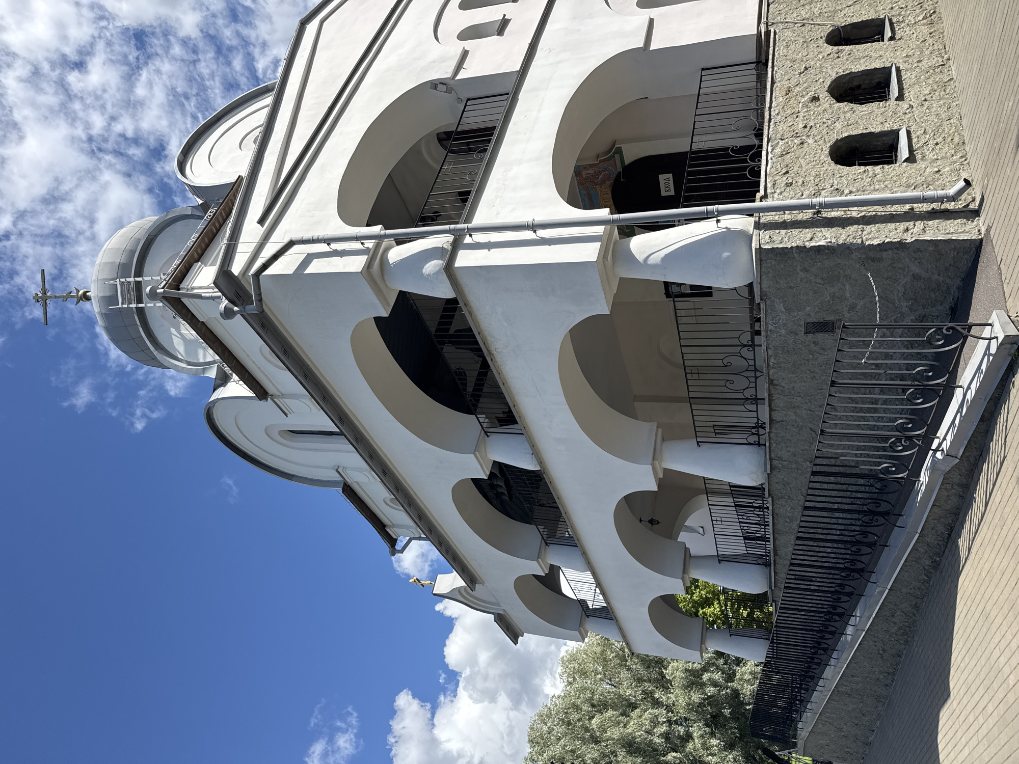 This is a medium-angle, exterior, eye-level shot of the Church of the Icon of the Mother of God of All Who Sorrow Joy located in Saint Petersburg, Russia. The church is made of white stucco and features a multi-tiered facade with a series of arched loggias. A domed roof with a cross topping it is visible at the highest point. The church is set on a stone foundation and is surrounded by a stone retaining wall. A ramp with a black metal railing leads up to one of the entrances. The sky is blue with scattered white clouds, and a lush green tree is visible on the left side of the frame. There are no people visible in the image. The lighting suggests it is a sunny day. The building has a predominantly neoclassical architectural style with some Byzantine influences.