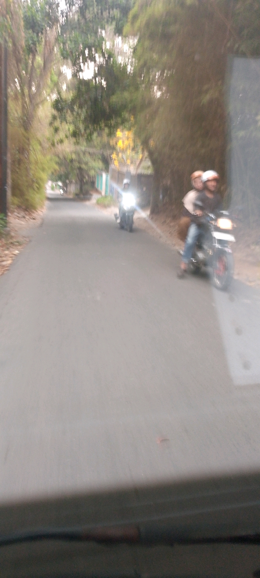 Two motorcycles are visible on a paved road in a setting with trees and foliage. The first motorcycle, closer to the viewer, has two individuals wearing helmets. The rider is in a dark shirt and blue jeans, and the passenger is behind them. The second motorcycle, further down the road, has a single rider wearing a helmet and appears to have its headlight on. The road is lined with trees on both sides, creating a shaded environment with some sunlight filtering through. The overall image is slightly blurred, suggesting movement. The scene likely takes place in a residential or rural area, given the presence of trees and the open road.