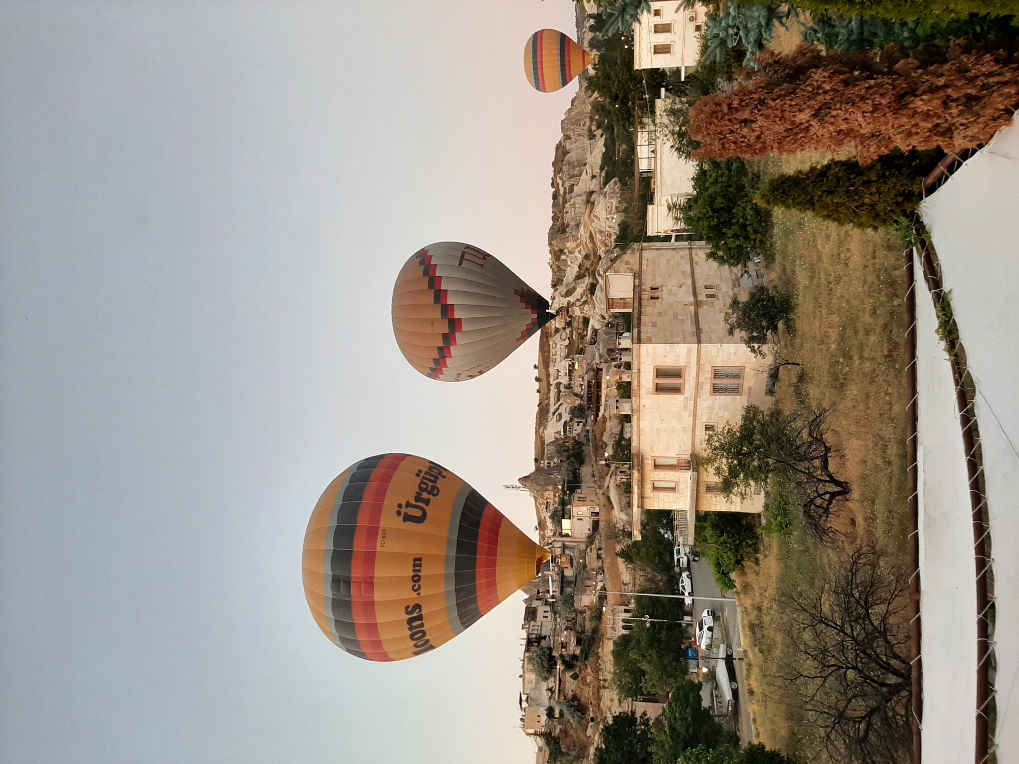The image depicts a landscape view in Göreme, Turkey, characterized by its unique geological formations and hot air balloon activities. Three hot air balloons are visible in the sky. The largest balloon in the foreground is predominantly orange with horizontal stripes of black and red, and it displays the text "loons.com Ürgüp". To its right, a second balloon is a muted gray with a pattern of small red and black squares. The third, smaller balloon on the far right, features horizontal stripes of orange and yellow.
Below the balloons, the scene transitions to a village built into and around a hillside. The architecture consists of structures made from light-colored stone, some appearing to be carved into the rock formations. Several buildings are clearly visible, including a multi-story house with a balcony in the mid-ground. Roads are present, with a few cars parked and driving, including a white car and what appears to be a trailer.
The natural environment is a mix of dry, grassy hillsides dotted with trees. Some trees are bare, showing intricate branches, while others are full of green foliage. In the foreground, a white canopy or awning is partially visible, suggesting a viewpoint or seating area. The overall lighting indicates either early morning or late evening, with the sky displaying a soft, pale gradient.