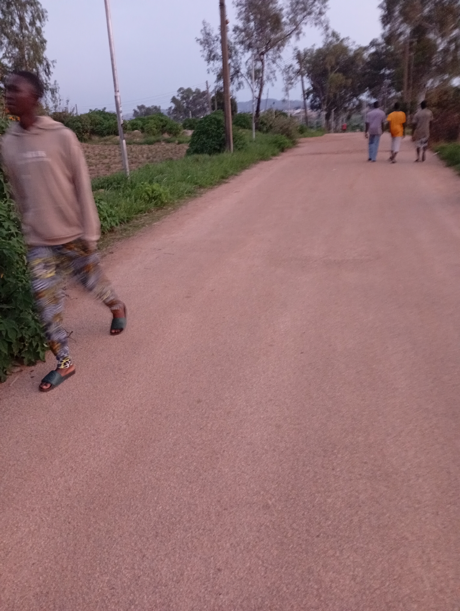 In the city of Jos, Nigeria, an individual wearing a light brown hooded sweatshirt and multi-colored patterned leggings is partially visible on the left side of a dusty, reddish-brown road. This person is seen from the waist down, with their left leg bent at the knee as they walk. They are wearing dark-colored slides on both feet. To the right of the frame, further down the road, three other individuals are walking away from the camera. The person in front is wearing a purple shirt and blue trousers, the middle person is in an orange shirt and white trousers, and the person behind them is wearing a grey shirt and patterned trousers. The environment suggests a rural or semi-rural setting, with sparse trees and vegetation lining the road. The sky is overcast, indicating an early morning or late evening.