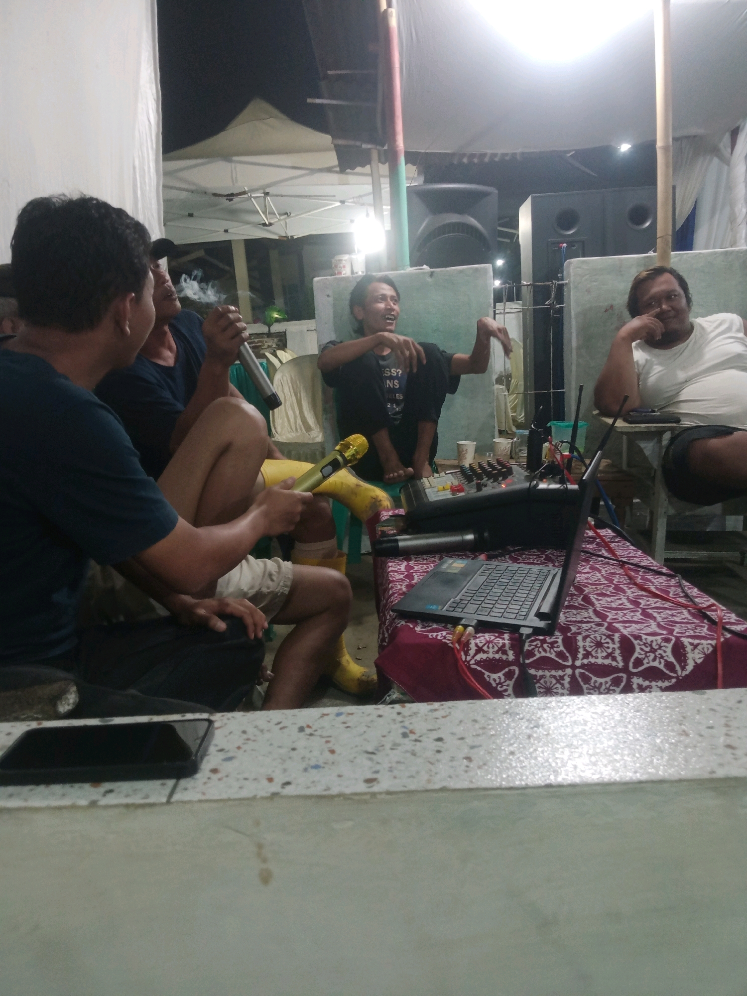 In Rejasari, Indonesia, a group of men gather outdoors under a makeshift tent, illuminated by artificial light. The scene appears to be a casual social event, possibly involving karaoke or music performance, given the presence of audio equipment. Three men are prominently visible, seated and engaged in conversation or activities related to the setup. One man, on the left, is holding a microphone and smoking, with smoke visibly rising. Another man, in the center, is leaning back with his arms outstretched, his expression suggesting a lively mood. On the right, a man in a white t-shirt is resting his chin on his hand, observing the scene. A table in the foreground is laden with a laptop, a sound mixer, and more microphones, all connected by wires. The surrounding environment includes large speakers and the tent structure, indicating an outdoor or semi-outdoor event. The overall atmosphere is informal and relaxed.