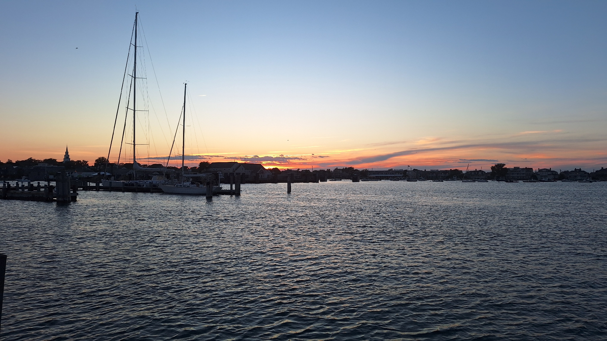 The image depicts a serene sunset over a harbor in Nantucket, Massachusetts, United States. In the foreground, dark, rippling water fills the lower half of the frame. Silhouetted against the twilight sky are several sailboats docked at a pier on the left. The tallest sailboat has two prominent masts. Further back, along the waterfront, a collection of buildings and trees form a town skyline. A church steeple is visible to the left of the tallest sailboat. The sky displays a gradient of colors, transitioning from a light blue at the top to warm oranges and pinks near the horizon, where the sun is setting. Wispy clouds stretch across the sky, catching the fading sunlight. The overall scene is tranquil and picturesque, characteristic of a coastal town at dusk.