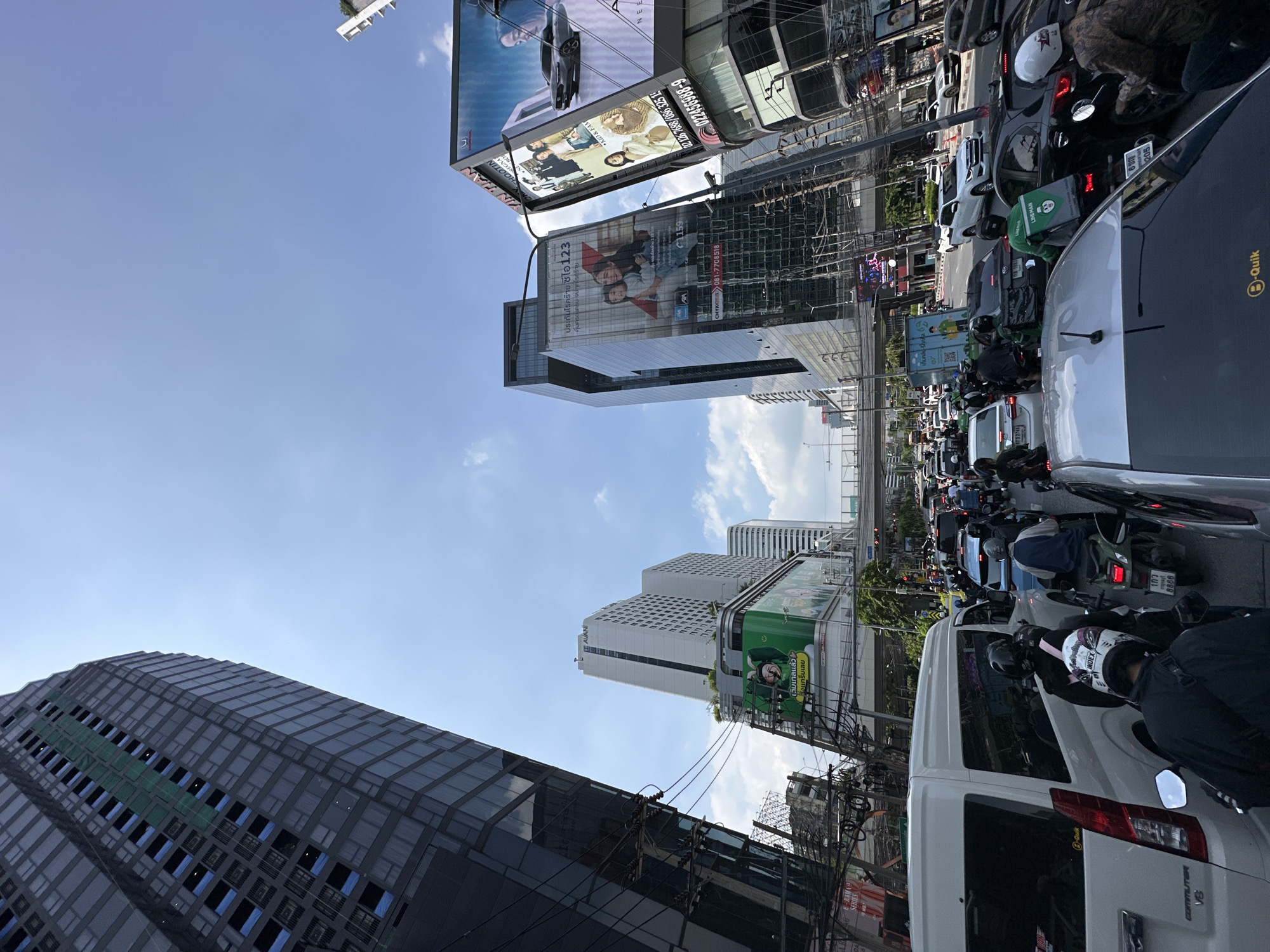 The image depicts a congested street scene in Bangkok, Thailand, framed by towering buildings adorned with large billboards. Traffic is heavy, with a mix of cars, vans, and a large number of motorcycles, many identifiable by their delivery service logos. The buildings are a mix of modern glass structures and more traditional high-rises, all framed against a blue sky with scattered clouds. Power lines are visible, adding to the urban clutter.
