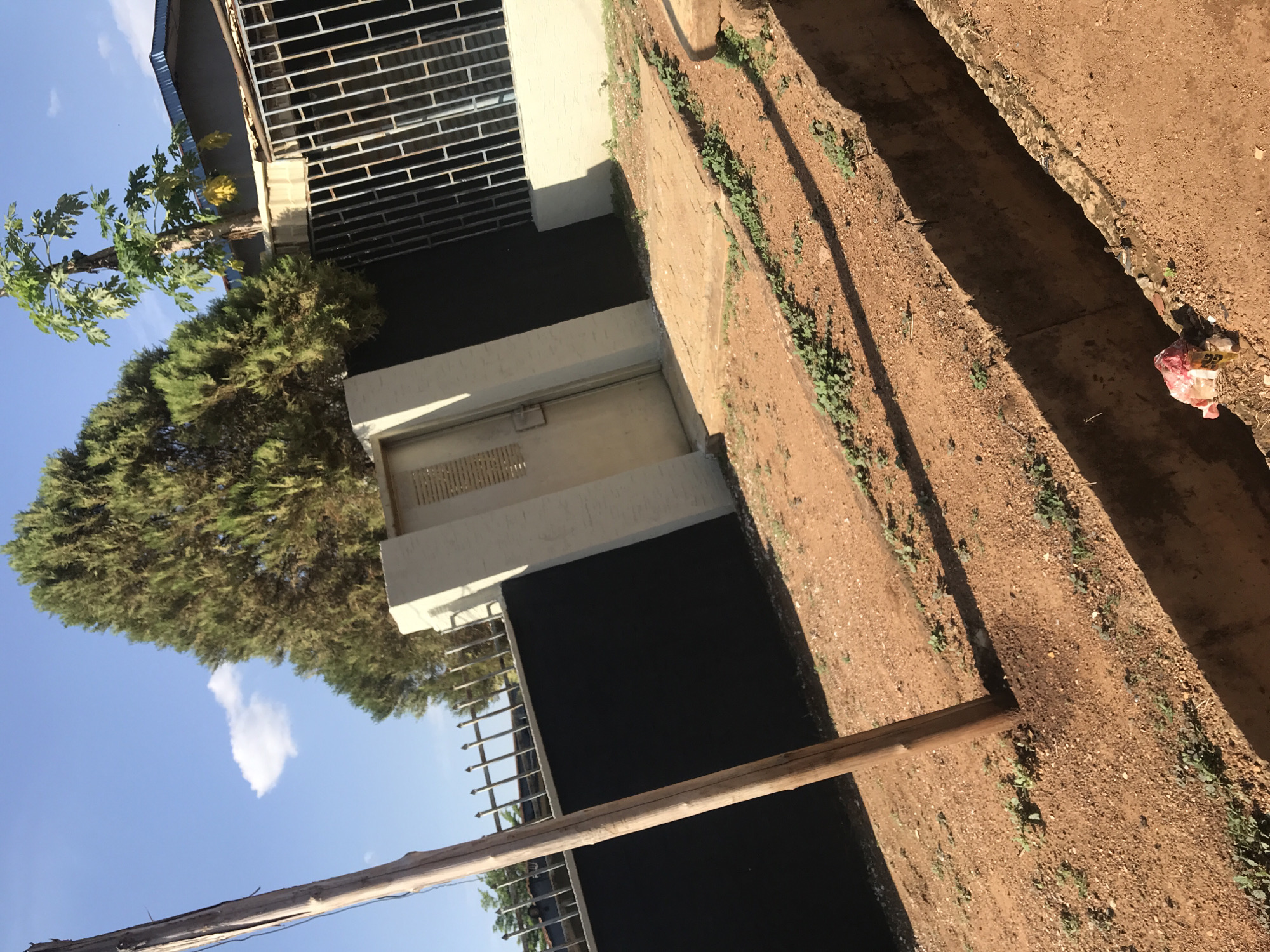 The scene is located in the Mandate Estate in Ilorin, Nigeria. The image depicts a building with a metal-barred window and a metal door. A large tree stands next to the building, reaching up into a partly cloudy sky. A wooden pole extends across the foreground. The ground is dirt and gravel, with some vegetation. A piece of trash lies on the ground.