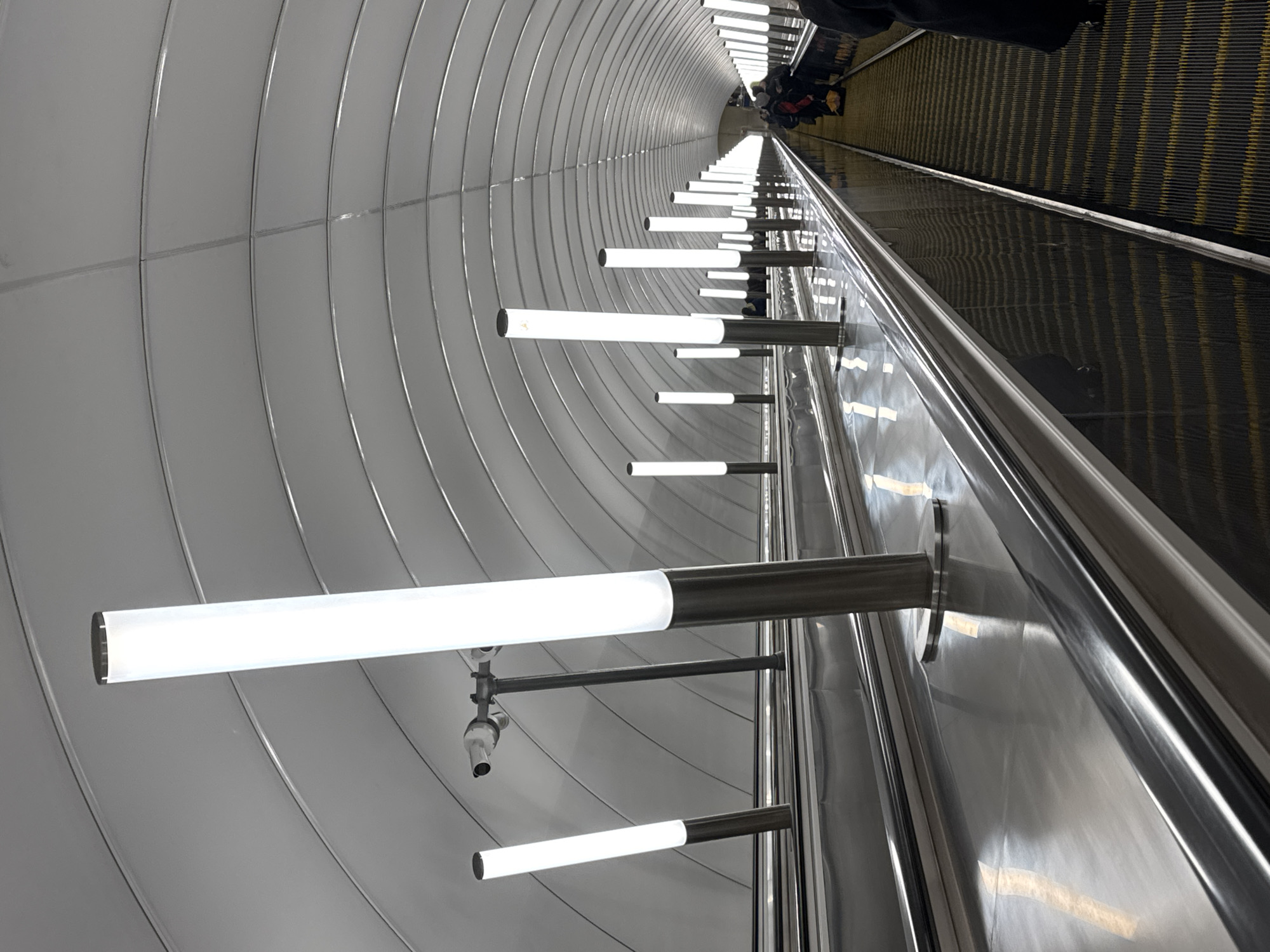 The image depicts a long escalator at Admiralteyskaya Metro Station in Saint Petersburg, Russia. The escalator ascends towards a tunnel with a curved, white-paneled ceiling illuminated by rows of horizontal, cylindrical lights. The escalator's steps are black with yellow stripes. A few people are visible at the top of the escalator. The metal handrails are polished, reflecting the lights. A surveillance camera is visible near one of the lights.