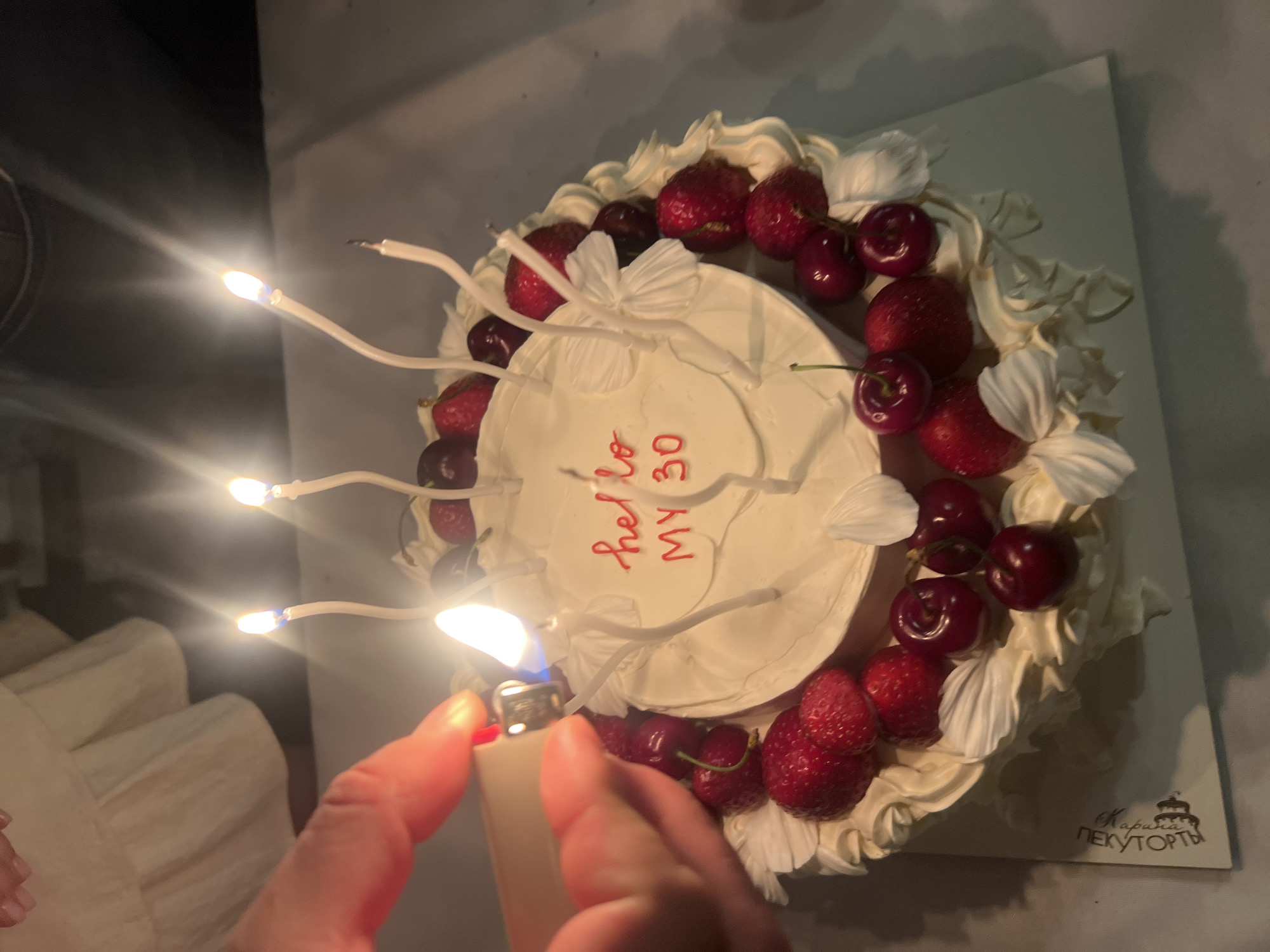 A close-up shot captures a hand holding a lighter to a lit birthday candle on a white cake. The cake is decorated with cream frosting, fresh strawberries, and cherries. Red frosting spells out "hello MY 30" on the top of the cake. Several unlit white candles are also visible around the cake. The background is dark and out of focus, with hints of a white ruffled garment. The bottom left corner of the image shows a white cake board with some text and a crest. The scene appears to be indoors, likely a celebratory setting. The specific location in Tomilino, Russia, cannot be determined from the visual information provided.