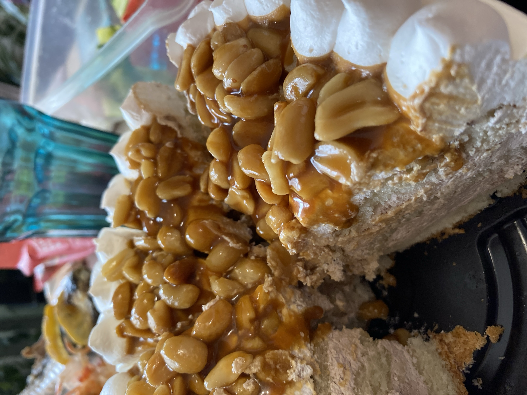 A close-up image shows a partially eaten cake. The cake has layers of white frosting, cake, and a topping of peanuts coated in caramel. The cake is on a black plastic plate or container. The location in the city of Kauchuk Stantsiya, Russia is unknown and cannot be determined from the image. There are no people visible in the image.