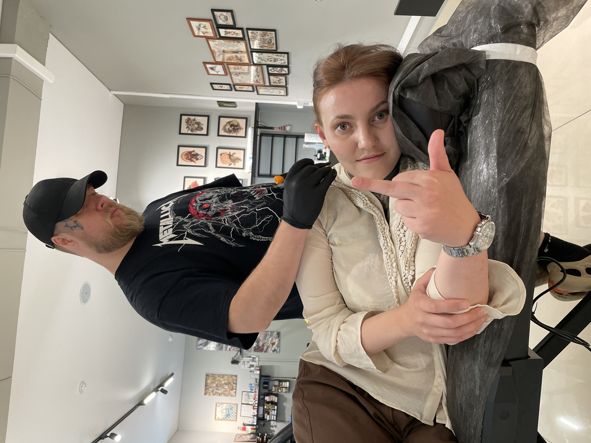 A man in a black baseball cap and a black t-shirt with a graphic print is tattooing a woman who is lying on her side on a tattoo table. The man is wearing black gloves and appears to be holding a tattoo gun. The woman is wearing a cream-colored blouse and brown pants. She is looking at the camera and has her middle finger raised. The background shows a tattoo studio with artwork displayed on the walls. The image is taken from a medium shot, focusing on the interaction between the tattoo artist and the client. The specific location within Izhevsk, Russia cannot be determined from the visual information provided.