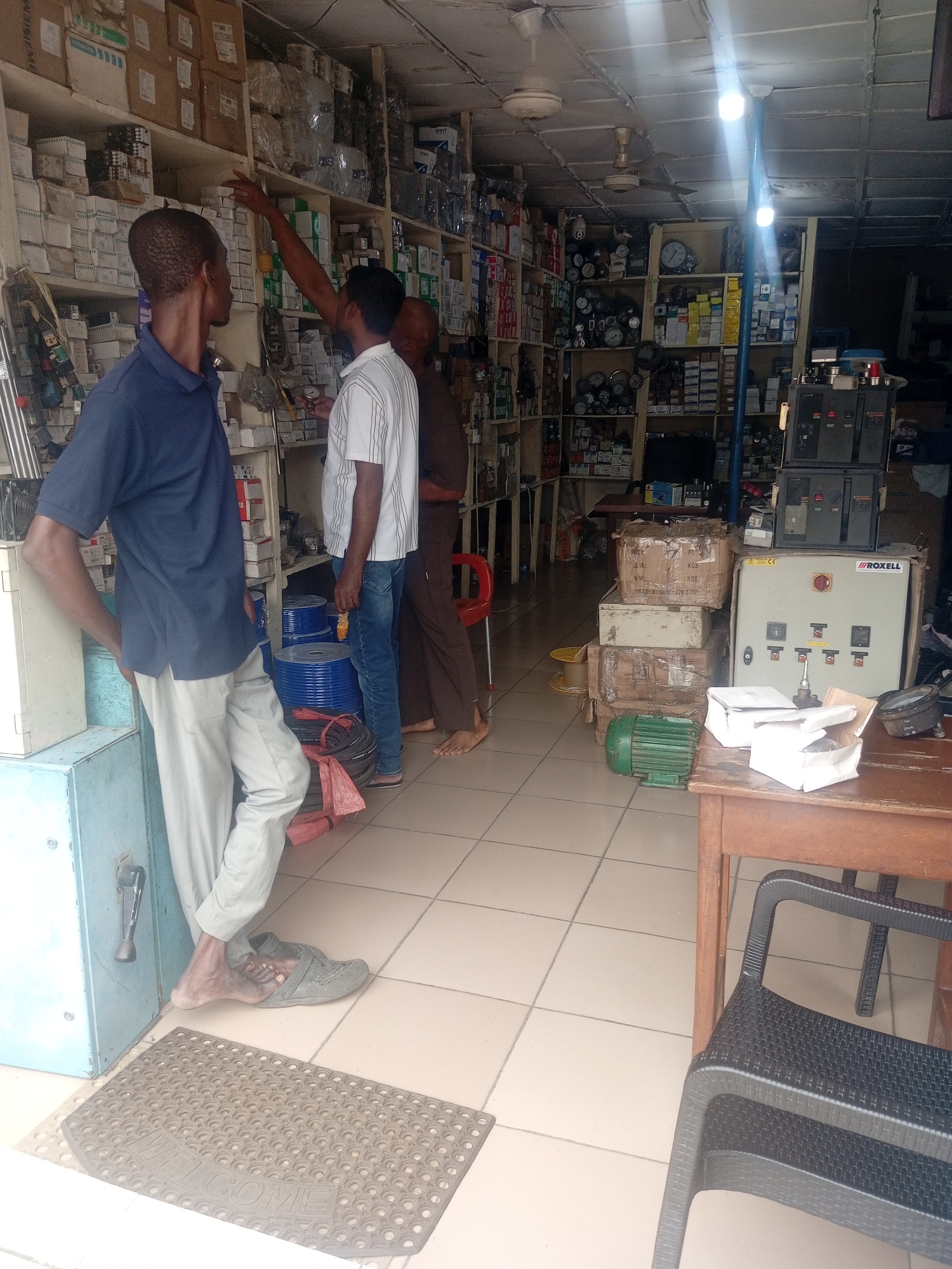 The image depicts the interior of an electrical supplies store, likely located in Sardauna, Nigeria, given the context. The store is densely packed with shelves filled with various electrical components, boxes, and equipment. Three men are present in the scene. One man, wearing a blue polo shirt and light-colored trousers, stands facing away from the camera, his right hand resting on a light blue metal cabinet. Another man, in a white and blue striped shirt and blue jeans, is reaching up towards a shelf, possibly selecting an item. A third man, wearing a brown shirt and trousers, stands behind him. In the foreground, a wooden table holds some boxed items and an electrical panel. Several large blue spools of wire are stacked on the floor, along with black electrical cables. A green electrical motor is also visible on the floor near the table. The environment appears to be a retail space, with tiled flooring and fluorescent lighting overhead.