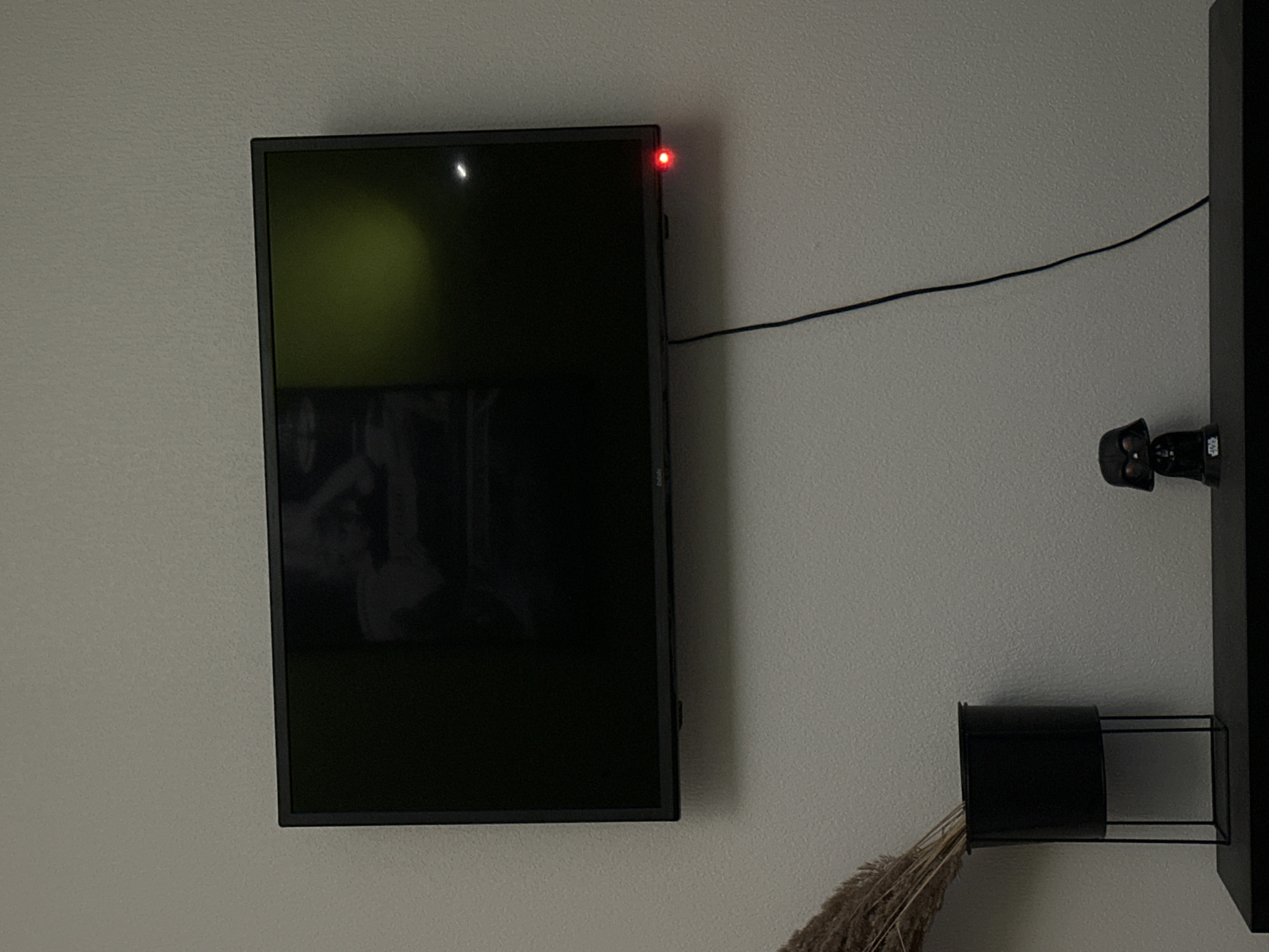 The image shows a dark-colored flat-screen television mounted on a light-colored textured wall. The television screen is black, reflecting a faint green and white light from an unseen source, and an image of a person can be vaguely seen on the screen. A small red indicator light is visible on the bottom right edge of the television. A black cable hangs down from the television. To the left of the television, a black metal planter on a black metal stand holds dried wheat or grass stalks. Below the television and planter, a black shelf is visible. On the shelf, to the left of center, is a small black figurine of Darth Vader from Star Wars. The overall scene appears to be an indoor domestic setting. There is no visible indication of the location being in the city of Unknown, Russia, or any specific geographic location.