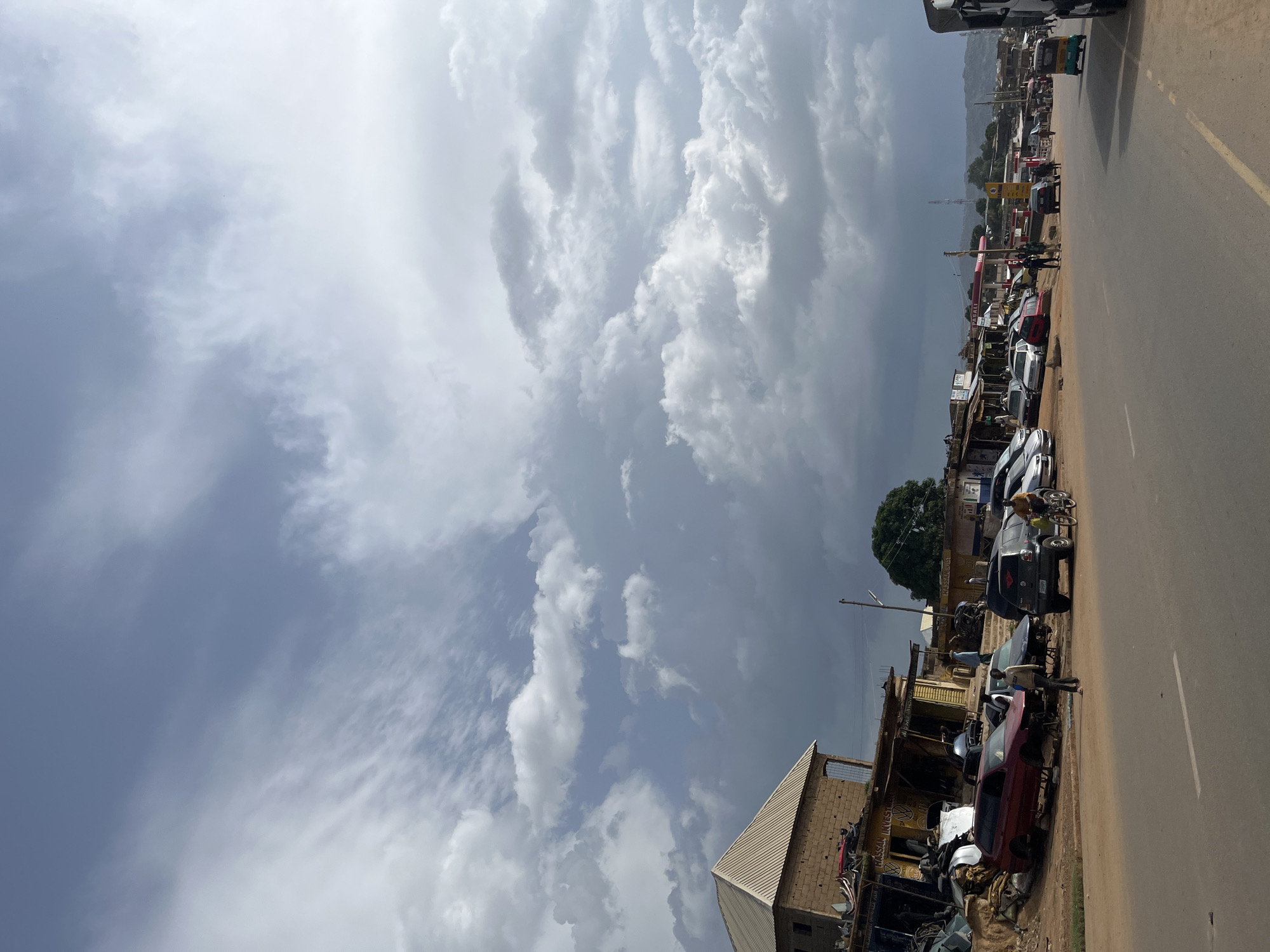 This is a full shot of a street scene in Jos, Nigeria, with a road in the foreground. On the right side of the road is a paved shoulder with several cars, a gas station, and various shops. On the left side of the road, there is a building with a corrugated metal roof, and several vehicles being dismantled for parts. People are seen walking and standing around the area, and some are interacting with the vehicles. A few of the vehicles appear to be in various states of disrepair, with some missing parts. The sky is filled with dramatic, grey clouds, suggesting a storm may be approaching. In the distance, a mountain range can be seen.