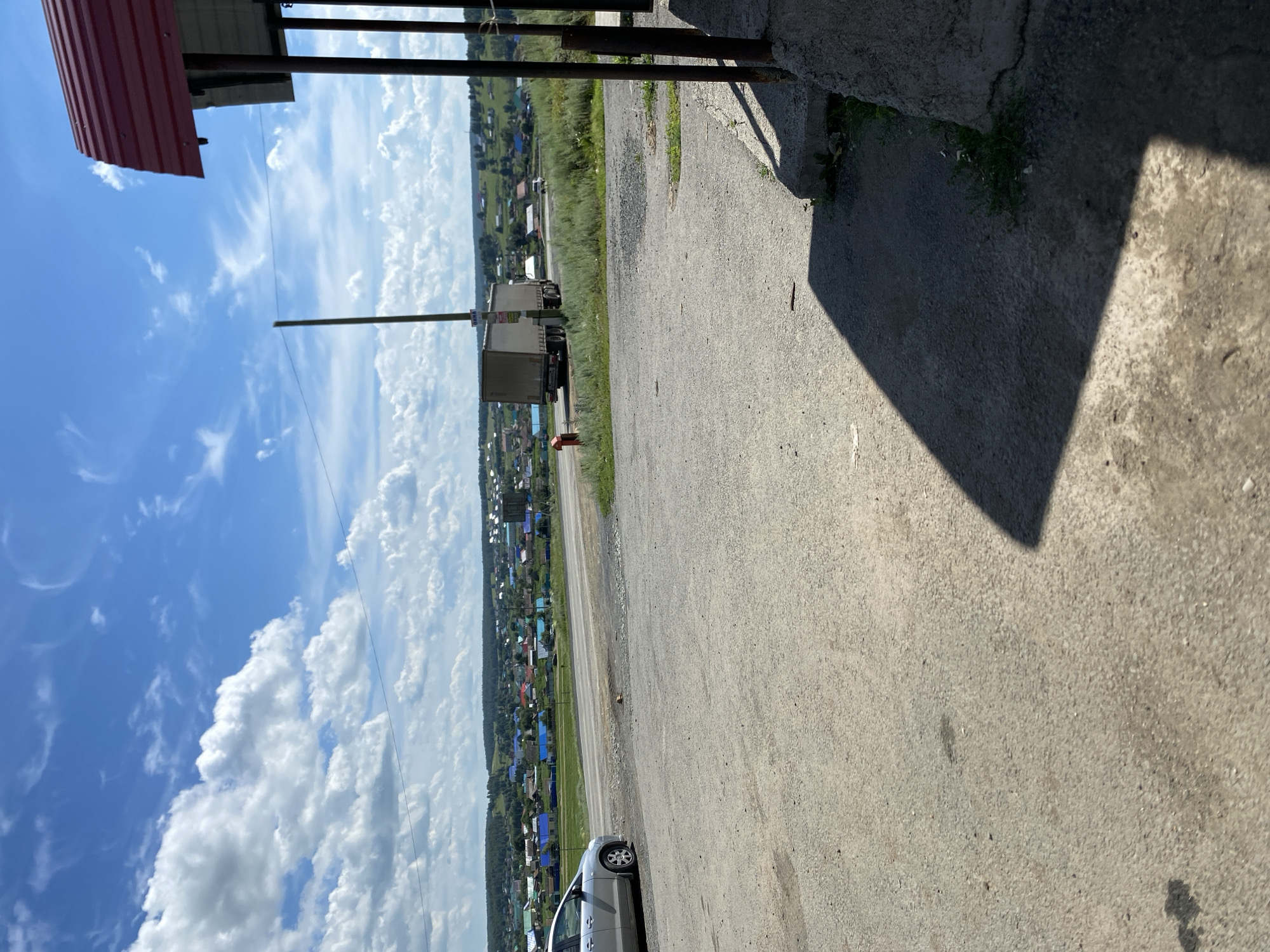 A bright, sunny day is depicted in Yuldybayevo, Russia, with a clear blue sky dotted with fluffy white clouds. In the foreground, a paved area transitions into a grassy verge. A silver car is parked on the left side of the paved area, facing left. Beyond the car, a road runs horizontally across the frame. In the middle ground, a large, beige truck is driving away from the camera on the road. Further in the distance, a cluster of houses with colorful roofs spreads across a gently rolling, green landscape, indicative of the village of Yuldybayevo. The right side of the image shows the corner of a building with a red metal roof and dark vertical supports. The overall scene is a rural roadside view with a village in the background.