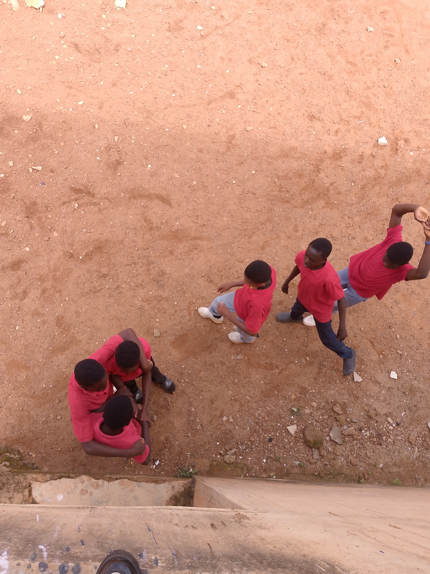 An overhead, high-angle medium shot shows five boys dressed in red shirts and dark pants. Three boys are positioned in the lower left of the frame, with two of them appearing to be in a playful wrestling or hugging embrace. The third boy is positioned to the left of them, his body turned away from the viewer, with his left arm raised and bent at the elbow. In the upper right quadrant of the frame, two boys stand further apart. One boy is facing away from the viewer and is in the process of raising his arms above his head, with his hands clasped. The second boy is facing towards the viewer and appears to be walking, with his left arm bent at the elbow and his right arm down by his side. They are standing on a surface of dirt and small stones. A concrete ledge is visible in the foreground, and a person's shoe is at the very bottom of the frame, suggesting the photo was taken from an elevated position.