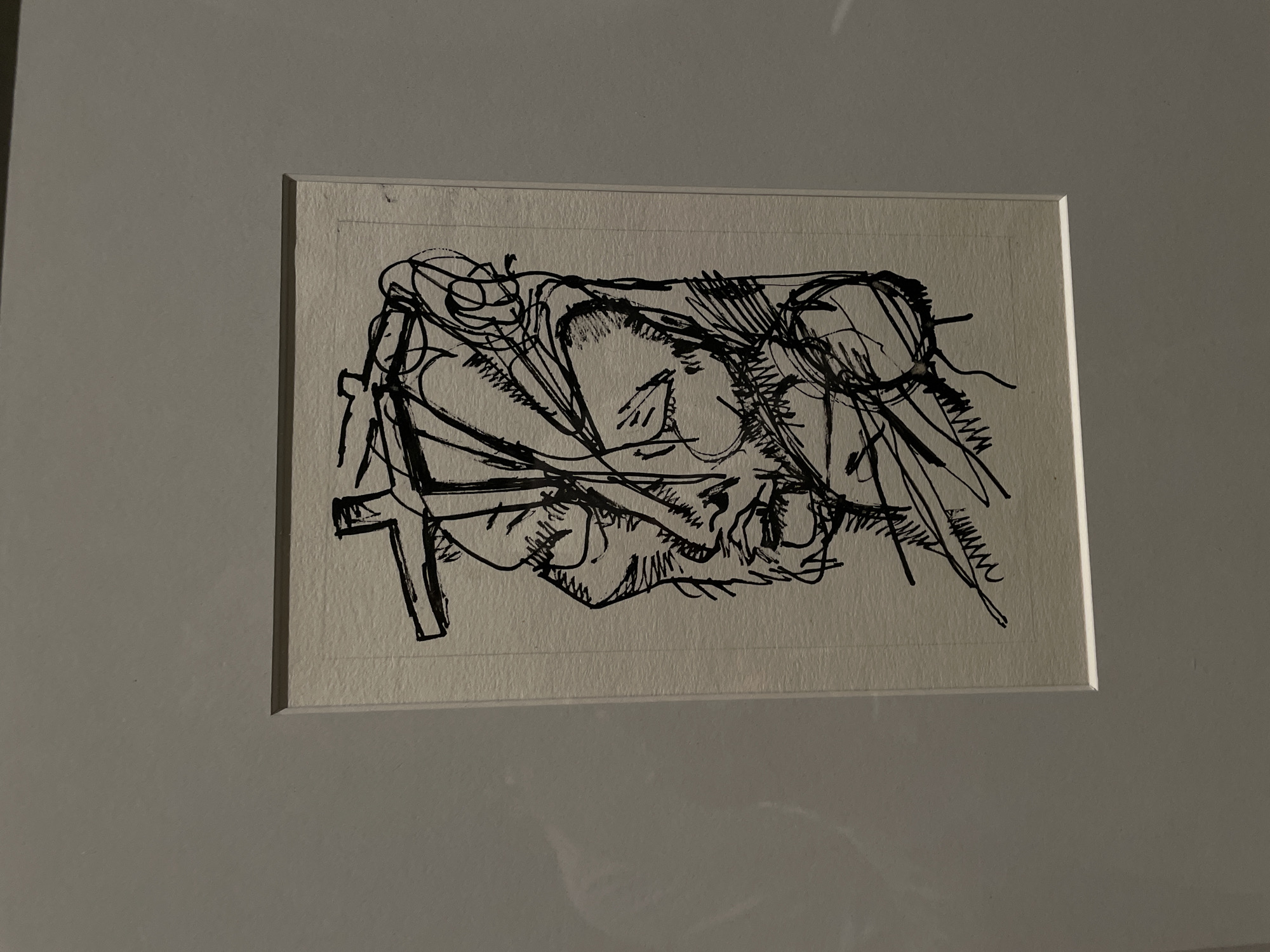 A framed, black ink drawing is displayed centrally on a plain, light grey wall. The artwork, rendered on textured light beige paper, features a highly stylized and abstract human head or face in left profile. It is characterized by thick, jagged, and swirling black lines that create distorted facial features, including an eye, nose, and mouth, along with an angular, cross-like structure positioned above the head. The drawing is bordered by a white mat, approximately 2-3 cm in width, which is enclosed within a thin, dark-colored frame. The environment indicates an indoor exhibition space. The specific location within Moscow, Russia, cannot be determined from the visual information in the image.