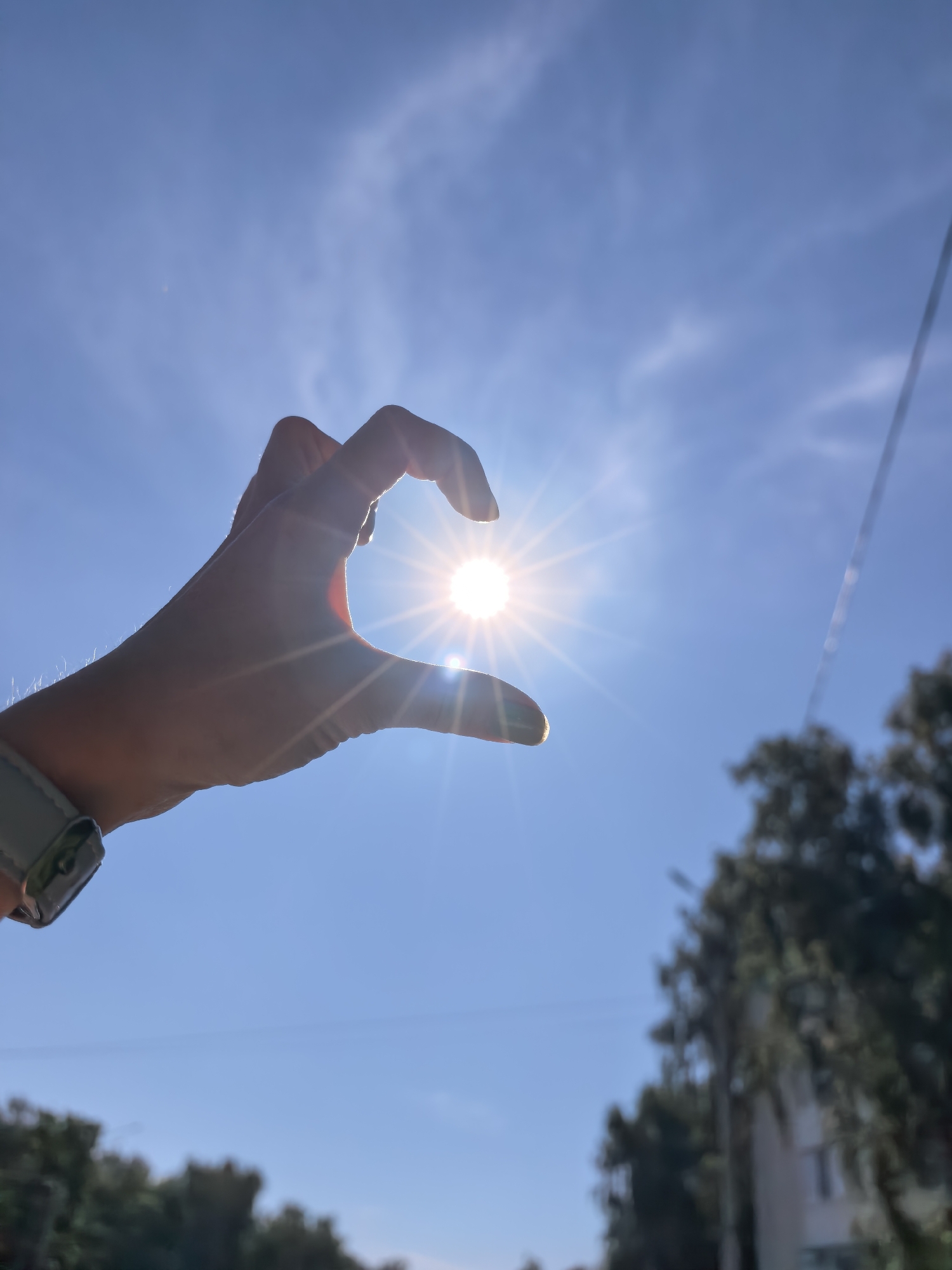 An outdoor, eye-level, medium shot captures a human hand silhouetted against a bright, sunny sky, forming a heart shape around the sun. The sun is a radiant starburst in the center of the frame, with visible rays emanating from it. The sky is a clear, vibrant blue with wispy white clouds scattered across it. A faint aerial wire is visible in the upper right quadrant of the image. In the background, the out-of-focus silhouettes of trees and a building suggest an urban environment. A smartwatch with a light-colored band is visible on the subject's wrist. The location is identified as Sterlitamak, Russia.