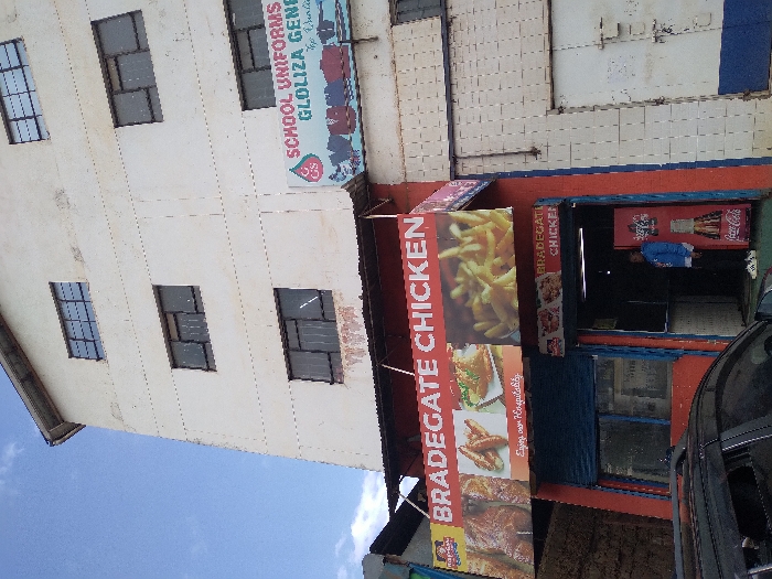This image captures a street view of a multi-story commercial building during the daytime. The building's exterior is painted a light cream or white color, with several windows visible on its upper floors. These windows have dark frames and appear to be fitted with security bars or a grid pattern.
On the upper part of the building, a large horizontal banner is displayed, advertising a business named "SCHOOL UNIFORMS GLOLIZA GEN". Below this text, there is a logo featuring the number "25" within a stylized shape, and possibly additional text that is difficult to read.
The ground floor of the building houses commercial establishments. On the left, a prominent vertical banner advertises "BRADEGATE CHICKEN". This banner showcases appetizing images of fried chicken, french fries, and other chicken dishes. Below these images, the phrase "Enjoy our Hospitality" is printed.
Adjacent to the large "BRADEGATE CHICKEN" banner, to the right, is the entrance to a shop, presumably also part of "BRADEGATE CHICKEN", as a smaller sign above its entrance also reads "BRADEGATE CHICKEN". The walls around this entrance are painted in bold red and blue sections. Inside the entrance, a Coca-Cola branded refrigerator is visible, and a person wearing a blue top is standing near it.
Part of a dark-colored vehicle, possibly black, is visible on the far right edge of the image, suggesting it is parked on the street in front of the shops. The sky in the background, visible in the upper left, is bright blue with some light clouds, indicating clear weather during daylight hours. The overall scene suggests a busy commercial area.