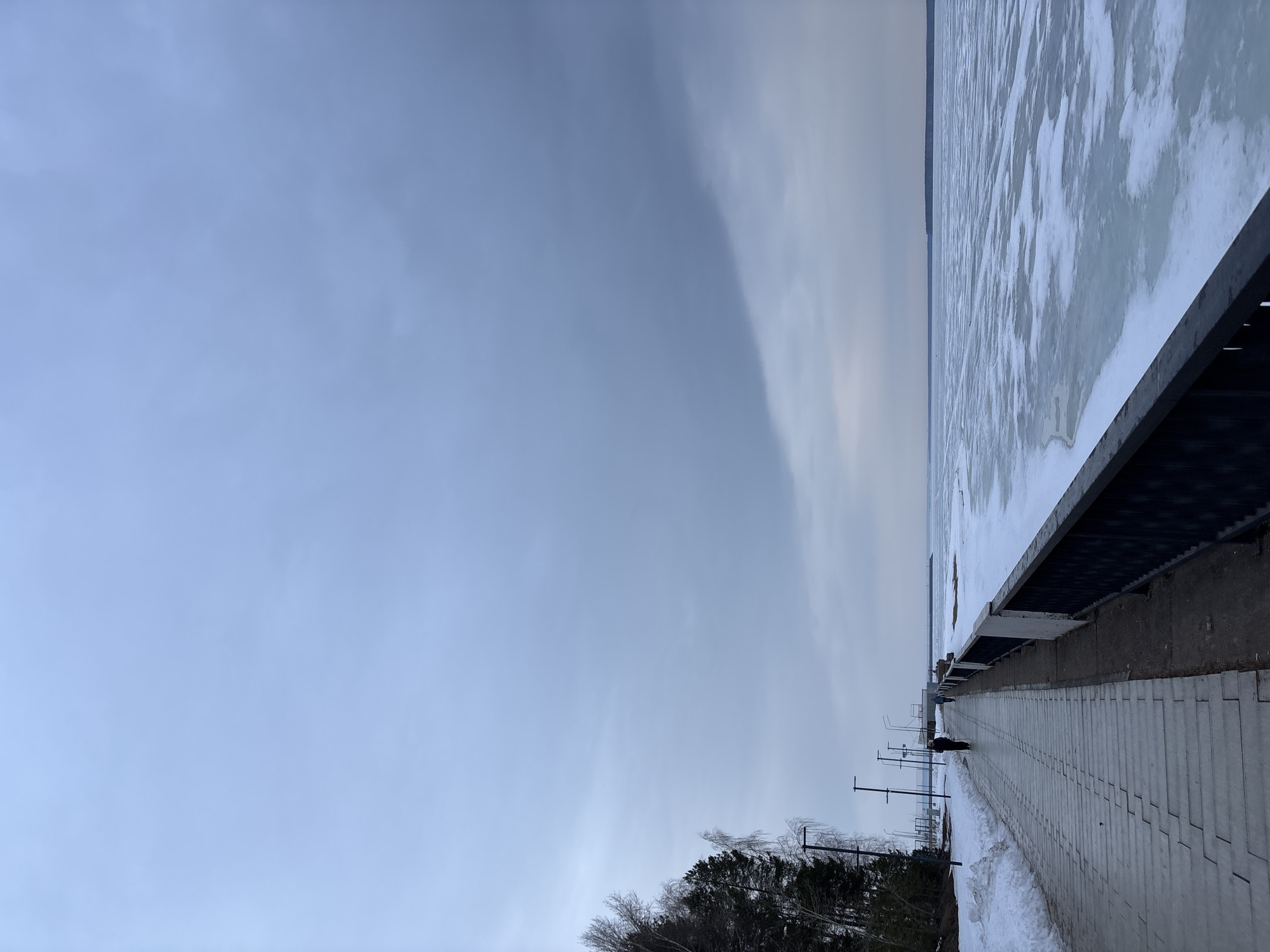 The image shows a landscape in the city of Chaikovsky, Russia. It's winter, and the view is dominated by a frozen river on the right side of the frame. A concrete embankment runs along the river, with a paved walkway made of grey stone tiles. The pathway extends into the distance, towards a single person walking. Along the walkway are evenly spaced lampposts. To the left of the walkway, there is snow and a sparse, low-lying forest or trees, seemingly covered in snow. The sky is overcast, a pale blue-grey, indicating a winter day.