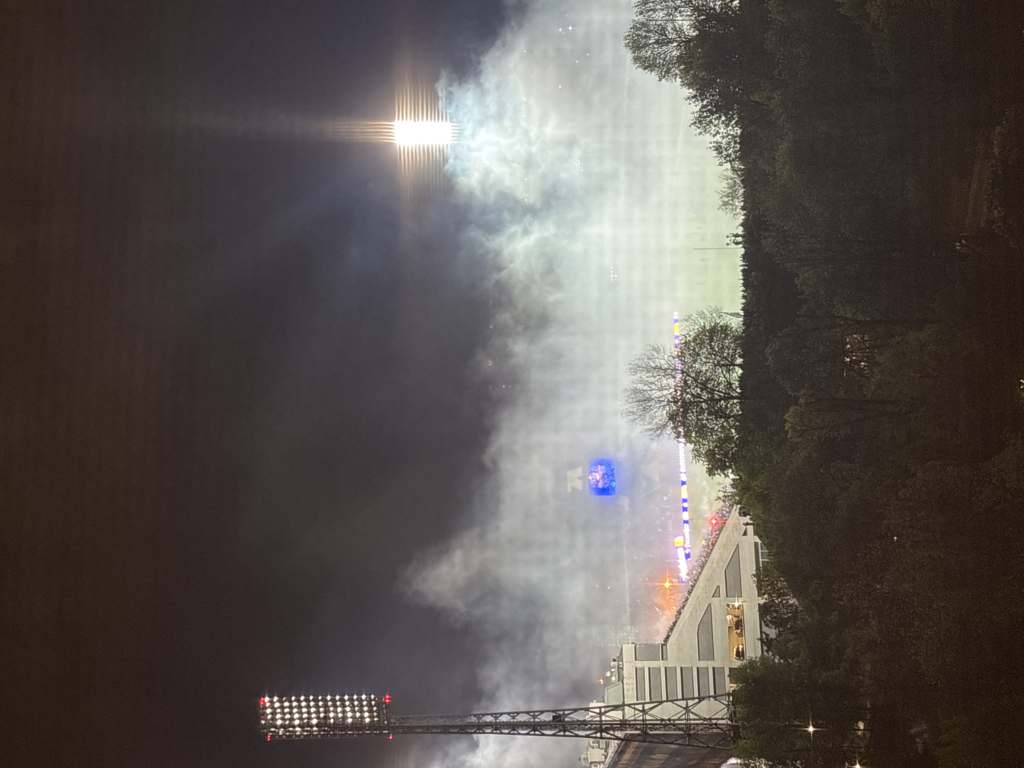 The image depicts a stadium at night. It shows the stadium lights illuminating a large cloud of smoke or fog over the field. The stadium appears to be crowded with people. The surrounding area includes trees and structures, possibly buildings or bridges. The specific location in the city of Unknown, Unknown cannot be determined from the image.