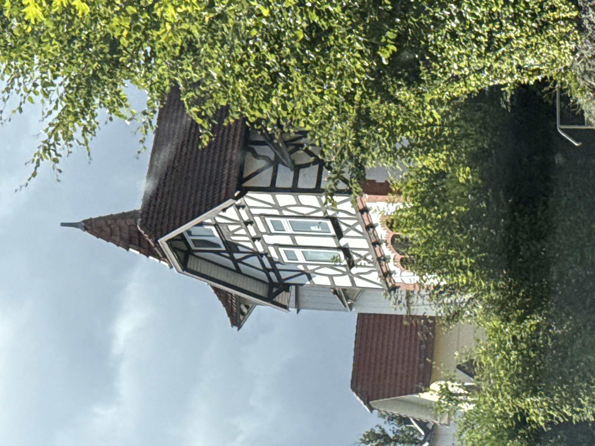 The image captures a view of a half-timbered house in Bad Sachsa, Germany, under a cloudy sky. The house features dark timber framing against a white plaster background. It has a steeply pitched, reddish-brown tiled roof, with a prominent pointed turret on the main structure and a smaller adjoining building with a similar roofline. Several windows with white frames are visible on the upper floors of the main house. Lush green trees and bushes partially obscure the lower sections of the building and the foreground. The overall scene suggests an exterior view on a day with overcast weather.