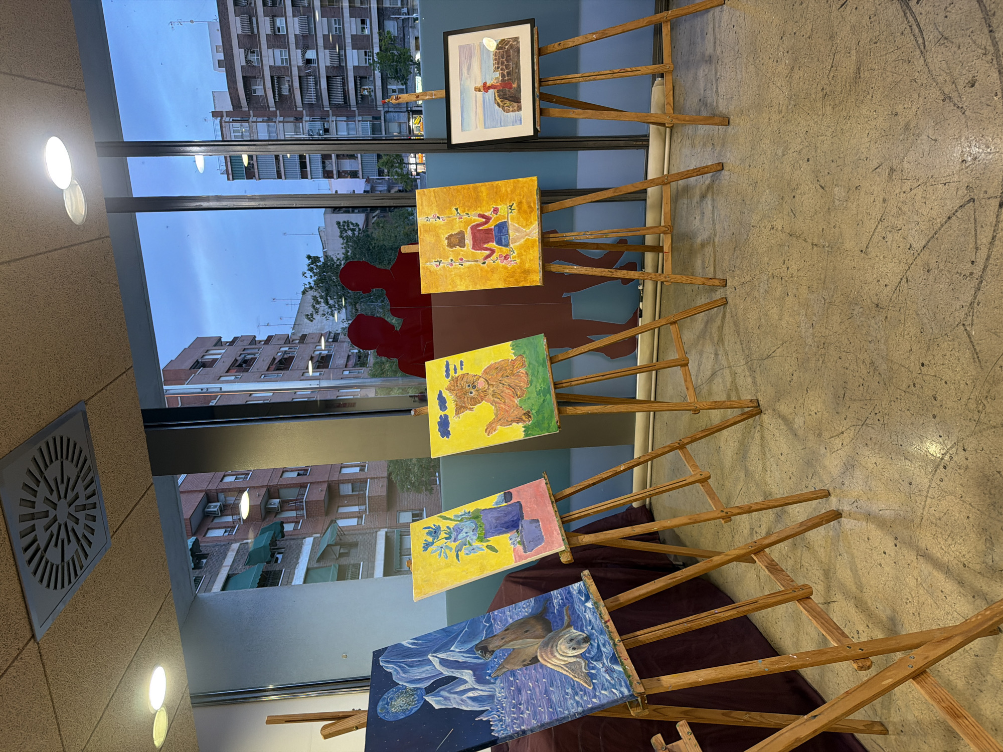 A collection of five paintings are displayed on wooden easels in front of a large window overlooking a city at dusk. The paintings, from left to right, depict: two seals on an icy landscape under a starry sky, a vase of blue flowers, a shaggy dog sitting on grass, a girl on a swing, and a person standing on a seawall. Silhouettes of two people are visible through the window. The environment appears to be an indoor exhibition space with a concrete floor and a suspended ceiling with air vents and light fixtures. The buildings outside the window are residential, with balconies and awnings.