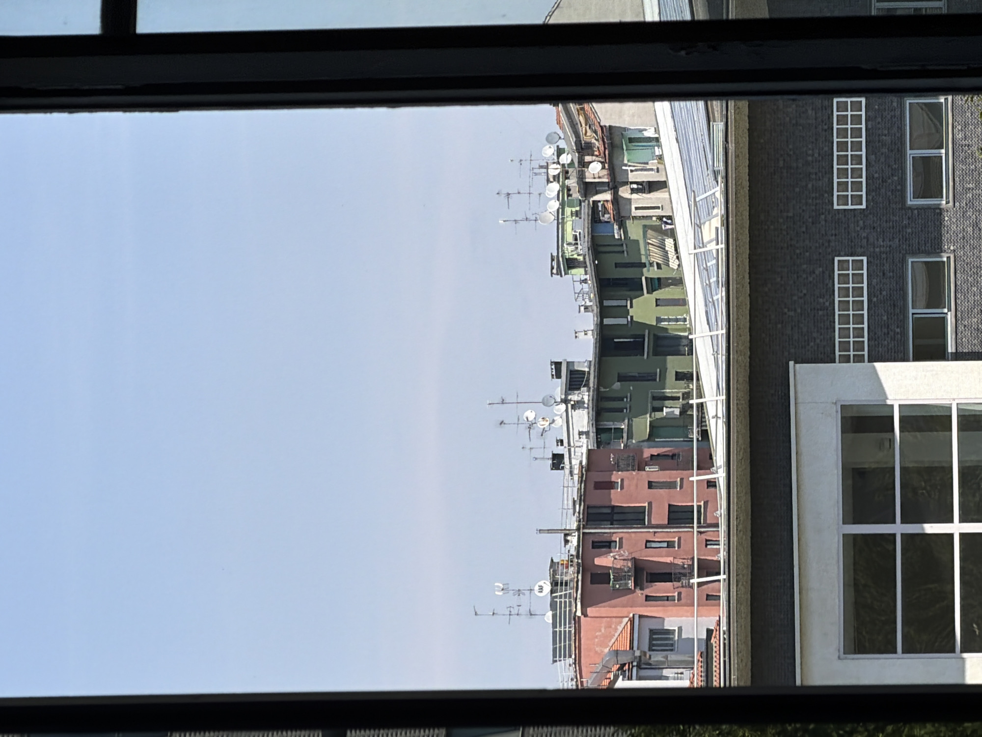 This image captures a view of buildings in Milan, Italy, seen through a window frame. The foreground is dominated by the dark, thick frame of the window, with a portion of a modern building visible below the rooftops. This lower building features a dark brick facade with horizontal bands of windows. Above this, the rooftops of several multi-story buildings are visible. The buildings themselves are varied in color and architectural style, with some painted in shades of pink and green, and others in muted tones. Many of the rooftops are adorned with numerous satellite dishes and antennas, suggesting a densely populated urban area with a need for telecommunication reception. The sky is a clear, pale blue, indicating fair weather. There are no people or vehicles directly visible in the scene. The overall impression is one of an urban landscape viewed from a residential or office building.