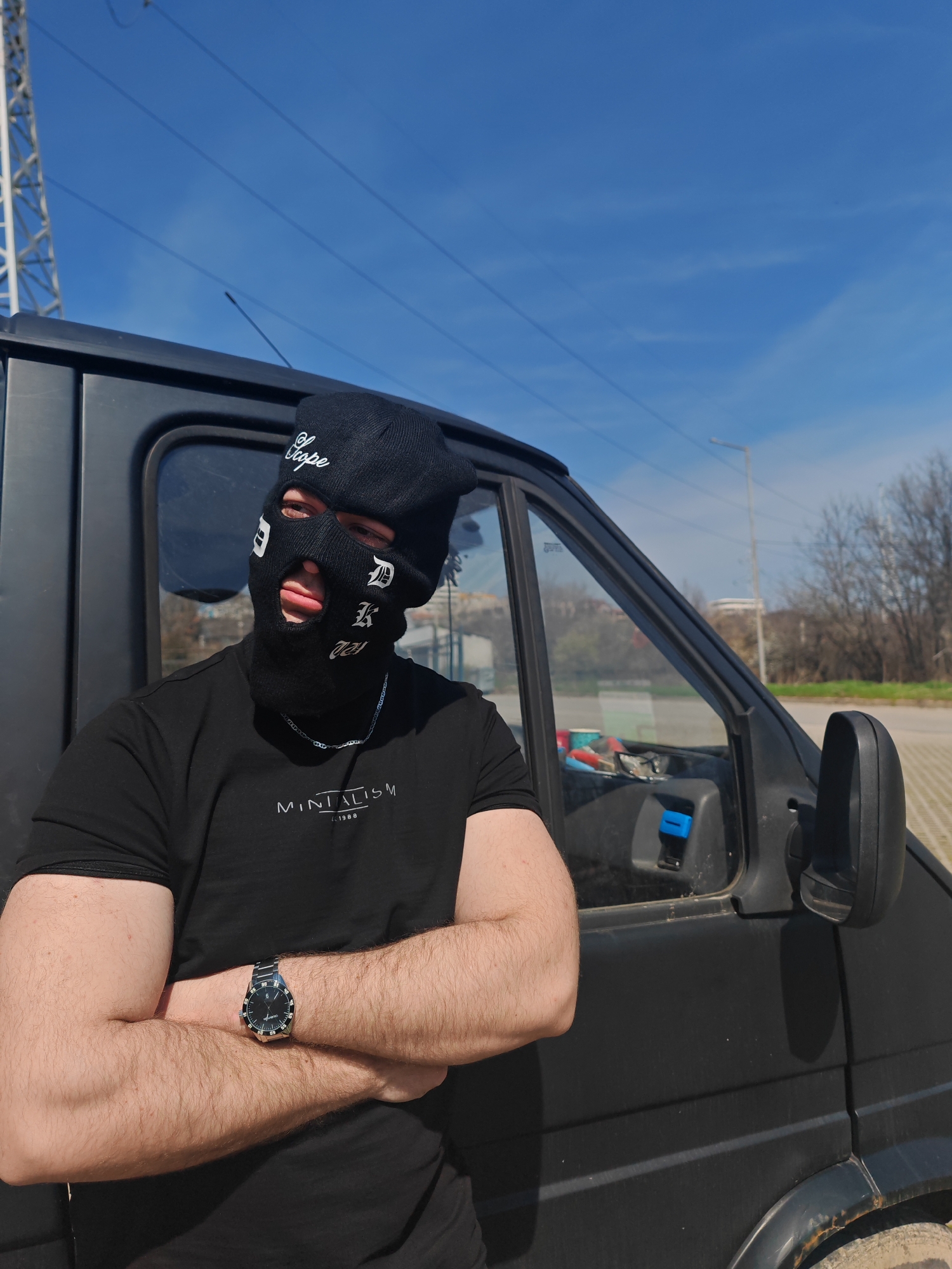 A male individual is depicted standing outdoors on a sunny day in what appears to be a parking or industrial area within Shumen, Bulgaria. He is positioned in front of the driver's side of a dark-colored panel van. The subject is wearing a black knitted balaclava with white text "Scope" on the forehead, "D" near the right eye, "K" near the left cheek, and "ZEU" below the mouth, revealing only a portion of his eyes and mouth. He wears a short-sleeved black t-shirt with "MINTALISM est. 1988" printed in white on the chest. A silver-colored chain necklace is visible. His arms are crossed at the chest, and a silver-colored wristwatch with a black face is worn on his left wrist. The van exhibits visible dirt, particularly around the wheel well. Through the van's driver-side window, tools or objects with red and blue components are discernible inside the vehicle. The background features a clear blue sky with some faint clouds, a metal lattice tower on the left, and a stand of bare trees or shrubs on the right. A street light pole is also visible in the mid-ground. The ground surface appears to be paved.