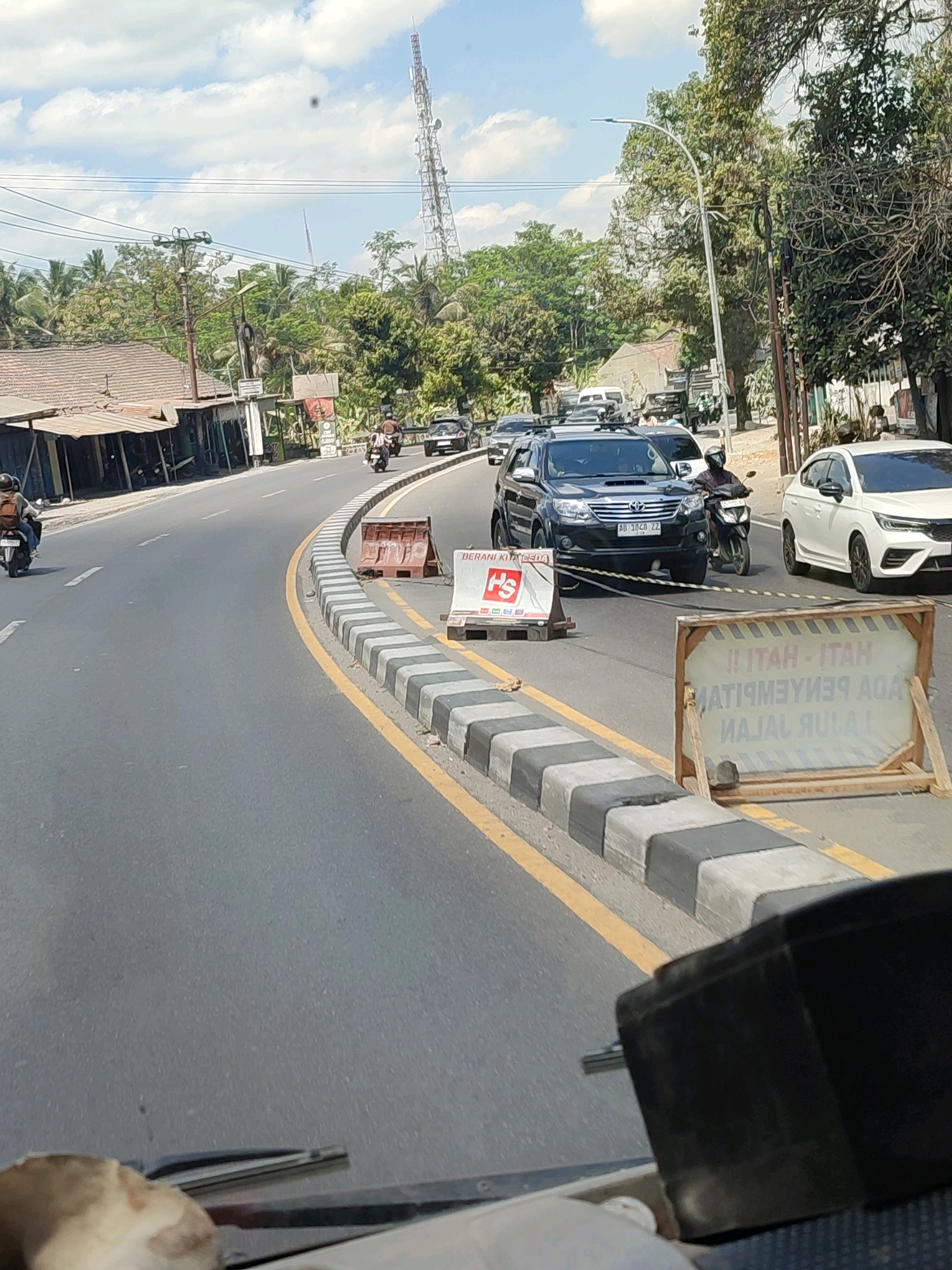 In Sucen, Indonesia, on a sunny day, a road with a yellow line marking the lane curves to the right. A concrete barrier with black and white stripes runs along the edge of the road. On the road, a black SUV with license plate AD 1020 ZZ is stopped, and a person on a motorcycle is driving past it. To the right of the SUV, a white car is also on the road. In the foreground, a temporary sign made of wood and fabric reads "!! HATI-HATI ADA PENYEMPITAN JALAN" in Indonesian, which translates to "!! BE CAREFUL THERE IS A ROAD NARROWING". Further down the road, another sign with the red "HS" logo is visible. The background shows a communication tower and lush green trees.