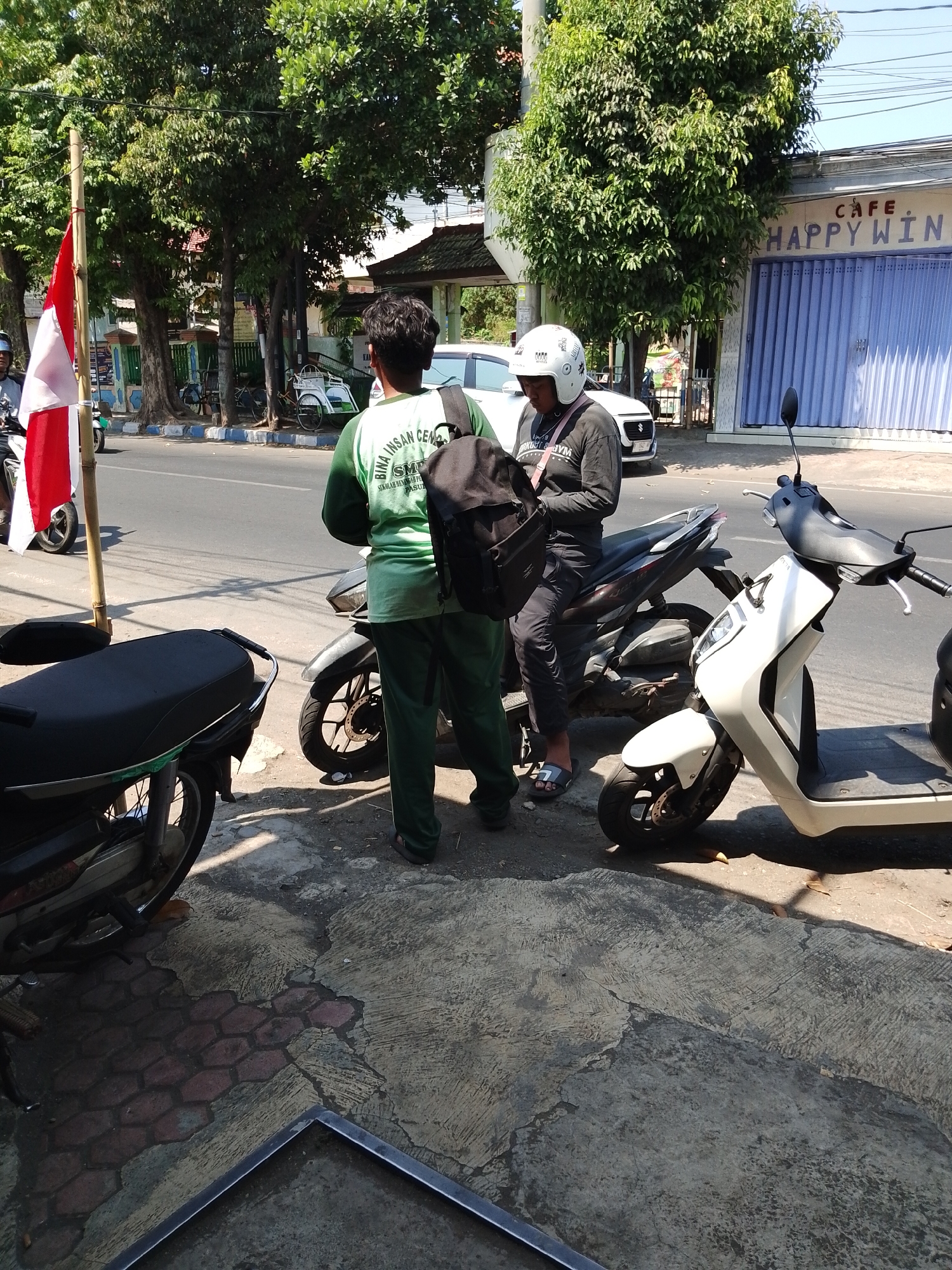 In Pasuruan, Indonesia, two individuals are standing next to scooters on a sidewalk. The person on the left is wearing a green long-sleeved shirt with text and a backpack. The person on the right, wearing a helmet and a black shirt, is seated on a scooter and looking at a phone. A red and white Indonesian flag is visible on a bamboo pole to the left. In the background, trees line the street, and a building with a sign reading "CAFE HAPPY WIN" is present. The ground is a mix of concrete and hexagonal paving stones.