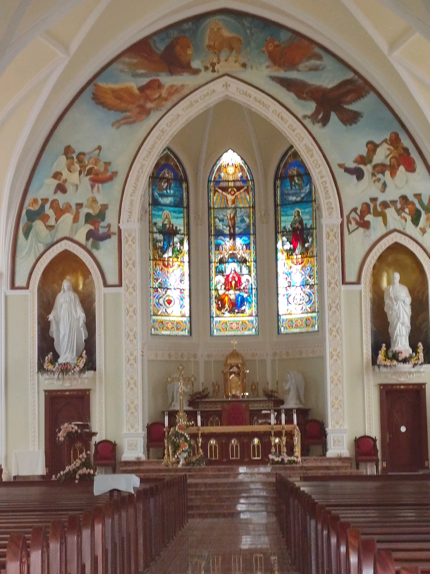 The image shows the interior of a Catholic church in Da Lat, Vietnam. The church features a high, arched ceiling with a mural depicting religious imagery, including lions, eagles, and figures that appear to be angels and people. Three stained-glass windows are prominently displayed in the center, depicting biblical scenes. A statue of what appears to be the Virgin Mary is housed in an alcove on the left, and another statue, possibly of an angel, is visible on the right. The altar area is elevated and decorated with floral arrangements and religious artifacts. Pews are arranged in rows facing the altar. The overall atmosphere is one of solemnity and religious devotion.