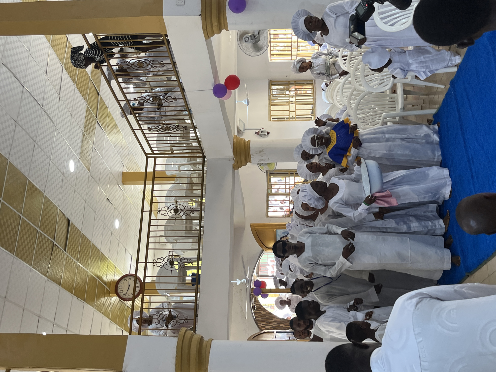 The image depicts an indoor gathering within a multi-level building, likely a community hall or religious establishment, located in an unknown city and country. The ceiling features a pattern of alternating white and gold textured acoustic tiles with recessed lighting.
On the upper level, a balcony with a decorative golden-bronze metal railing runs across the scene. To the far left, a person in a white robe stands behind the railing, near a wall clock displaying approximately 1:55. Several other individuals in white attire are seated behind the railing further right. One person, wearing a black and white striped top and a patterned headwrap, leans over the railing, observing the activity below. The balcony is supported by white fluted columns with gold capitals.
The main floor features a prominent blue carpet runner forming a central aisle. A large group of people, including adults and children, are gathered, most dressed in matching white robes or gowns. Many women wear white, ruffled head coverings, while some men wear white caps.
In the center foreground of the main floor, a person's back is visible, wearing a white textured garment, facing the main group. To the right of the blue runner, a woman in a white gown and head covering holds a camera with an external flash. A small child, also in white robes and a cap, stands facing away on the blue runner. To the left of the runner, a woman in a blue and yellow patterned top over a white gown, with white-framed sunglasses and a white head covering, smiles broadly and raises her hands slightly. Next to her, another woman in white holds a white basin and a pink cloth. Many individuals in the main group appear to be barefoot on the blue runner.
Further back on the main floor, a man wearing sunglasses and a white robe stands near the center. Other men in white robes, some with patterned fabric details or beaded necklaces, are present. An open arched doorway in the background reveals more people and exterior light. Walls are white, and several barred windows are visible. White plastic chairs are stacked or arranged on the right side of the main floor. Two clusters of balloons (red, pink, purple) are suspended from the ceiling. Multiple ceiling fans and wall-mounted oscillating fans are also visible.