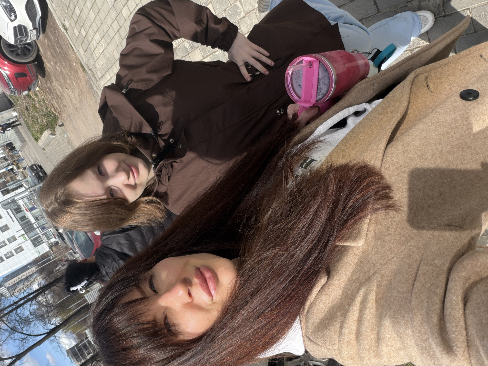 Two young women lie on a paved surface with cars and buildings in the background, indicative of an urban environment. The woman on the left wears a brown jacket and light blue pants. The woman on the right wears a beige coat and has dark brown hair. A pink drinking container is visible near the woman in the brown jacket. The presence of specific architectural features and landmarks suggests that the location is Perm, Russia.