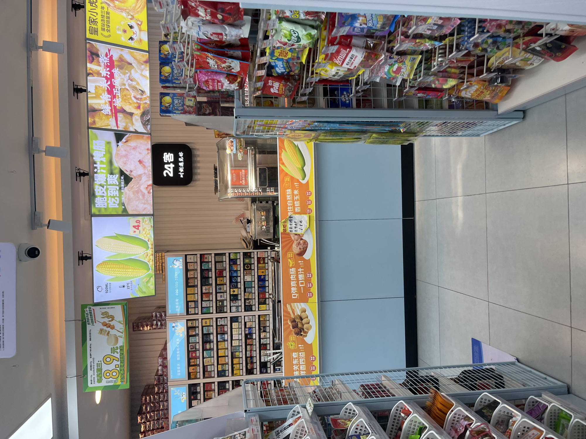 The image shows the interior of a convenience store, likely a 24-hour operation, in the Yuzhong District of China. The store is brightly lit, with fluorescent lighting overhead and illuminated display signs. The scene is organized and clean, with merchandise neatly arranged on shelves and in displays.
In the foreground, to the left, are shelves stocked with various food items and snacks, presented in white plastic baskets. To the right, a metal display rack is filled with numerous colorful packages of candy and snacks. The shelves are meticulously organized, indicating a well-managed retail environment.
The middle ground of the image features a counter area with a light blue facade. Above the counter, several illuminated signs display promotions for food items. One sign shows a price of 8.9 yuan along with images of skewers. Another sign features two ears of corn with a price of 4.0 yuan. There are also signs advertising roasted chicken and sausages. Behind the counter, a glass-fronted display case contains more food items, and a coffee machine or similar beverage dispenser is visible. Behind the counter, shelves are stacked with rows of cigarette packs.
The background of the image shows a wall with more shelves of cigarette packs, suggesting this is a prominent item sold at the store. A security camera is mounted on the ceiling. The floor is tiled with a light-colored material. The overall impression is that of a modern, well-stocked convenience store catering to a variety of customer needs. No people are visible in the image, suggesting the store is either empty or the photo was taken at a time when customer traffic is low.