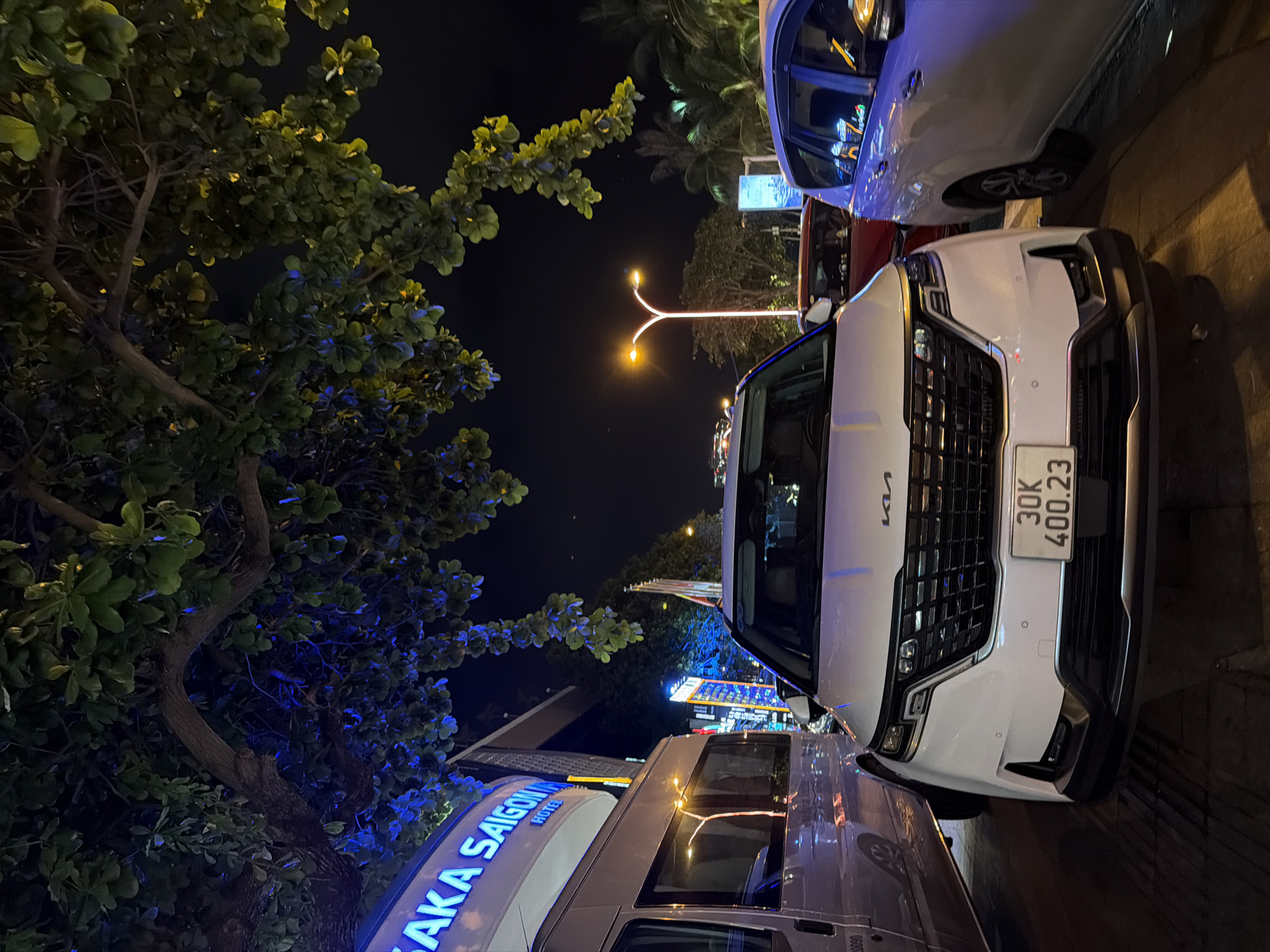 The image depicts a street scene at night in Nha Trang, Vietnam. In the foreground, a white Kia Carnival minivan is parked on a paved surface. The vehicle has a license plate that reads "30K 400.23". To the left of the Kia, a white van is parked, and to the right, a light purple minivan is visible.
Above the vehicles, a large tree with dark green leaves hangs over the street, illuminated by streetlights and building lights. The background shows a building with the words "SAKA SAIGON" in large, blue neon letters, indicating it is likely a hotel. Further in the background, streetlights and other illuminated signs are visible, suggesting a commercial or busy urban area. Palm trees can also be seen in the distance. The overall atmosphere is one of a vibrant city at night.