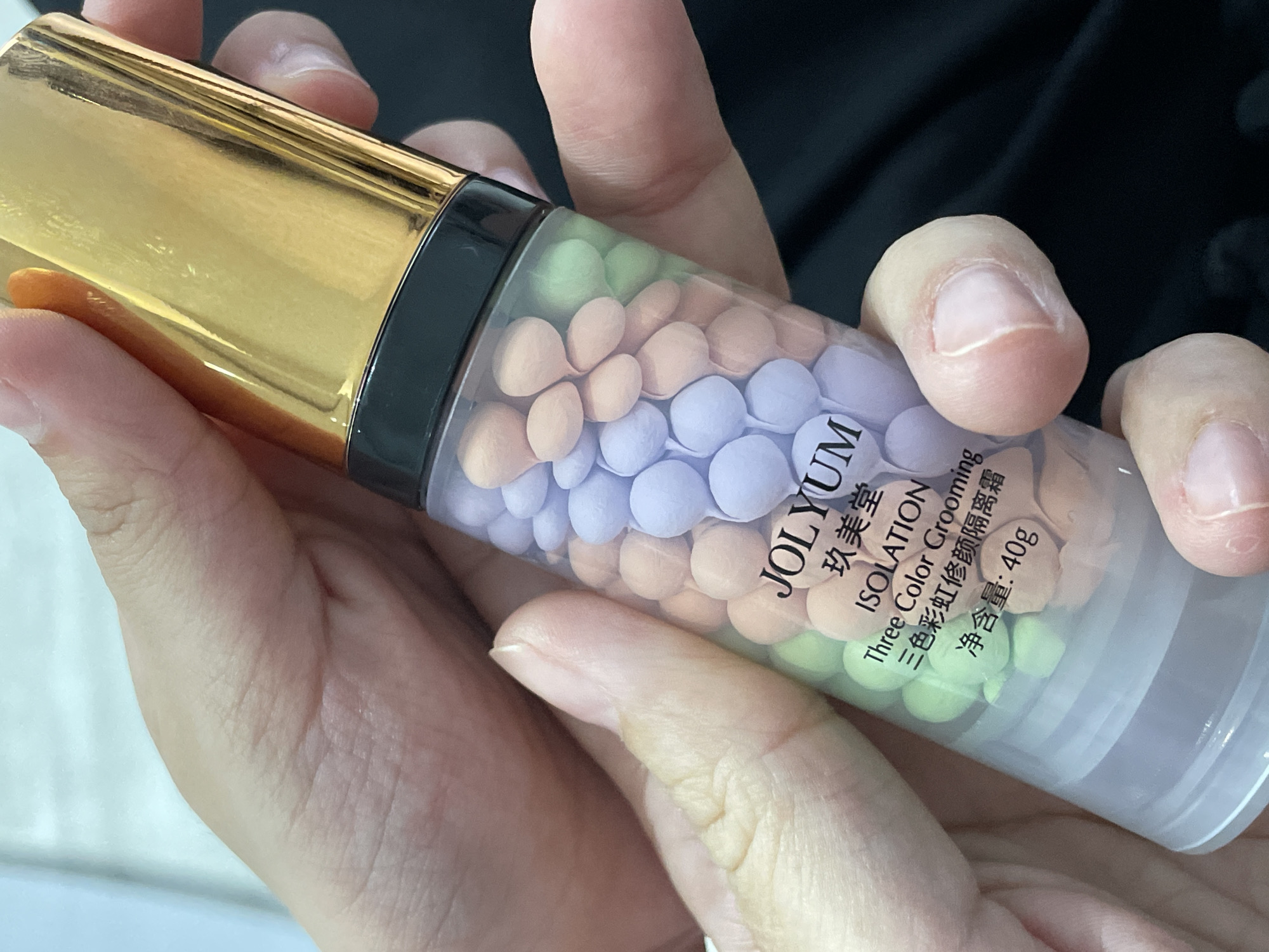 A close-up shot captures a person's hands holding a cylindrical cosmetic bottle. The bottle features a shiny, reflective golden cap and a transparent body filled with numerous small, spherical beads in three distinct pastel colors: light green, light orange/peach, and light purple.
The label on the bottle is clearly visible, displaying the brand name "JOLYUM" and its Chinese equivalent "玖美堂". Below this, the text "ISOLATION" and "Three Color Grooming" are present, alongside their Chinese translation "三色彩虹修颜隔离霜". The net weight, "净含量:40g", is also printed on the label.
The hands holding the bottle show visible knuckles and fingernails, suggesting an adult's hands. The background is softly blurred, appearing as a dark, possibly fabric, surface on the right and a lighter, indistinct area on the left, indicating an indoor setting. The lighting is bright, emphasizing the product. The person appears to be holding the product for display or examination. This scene is located in Izhevsk, Russia.