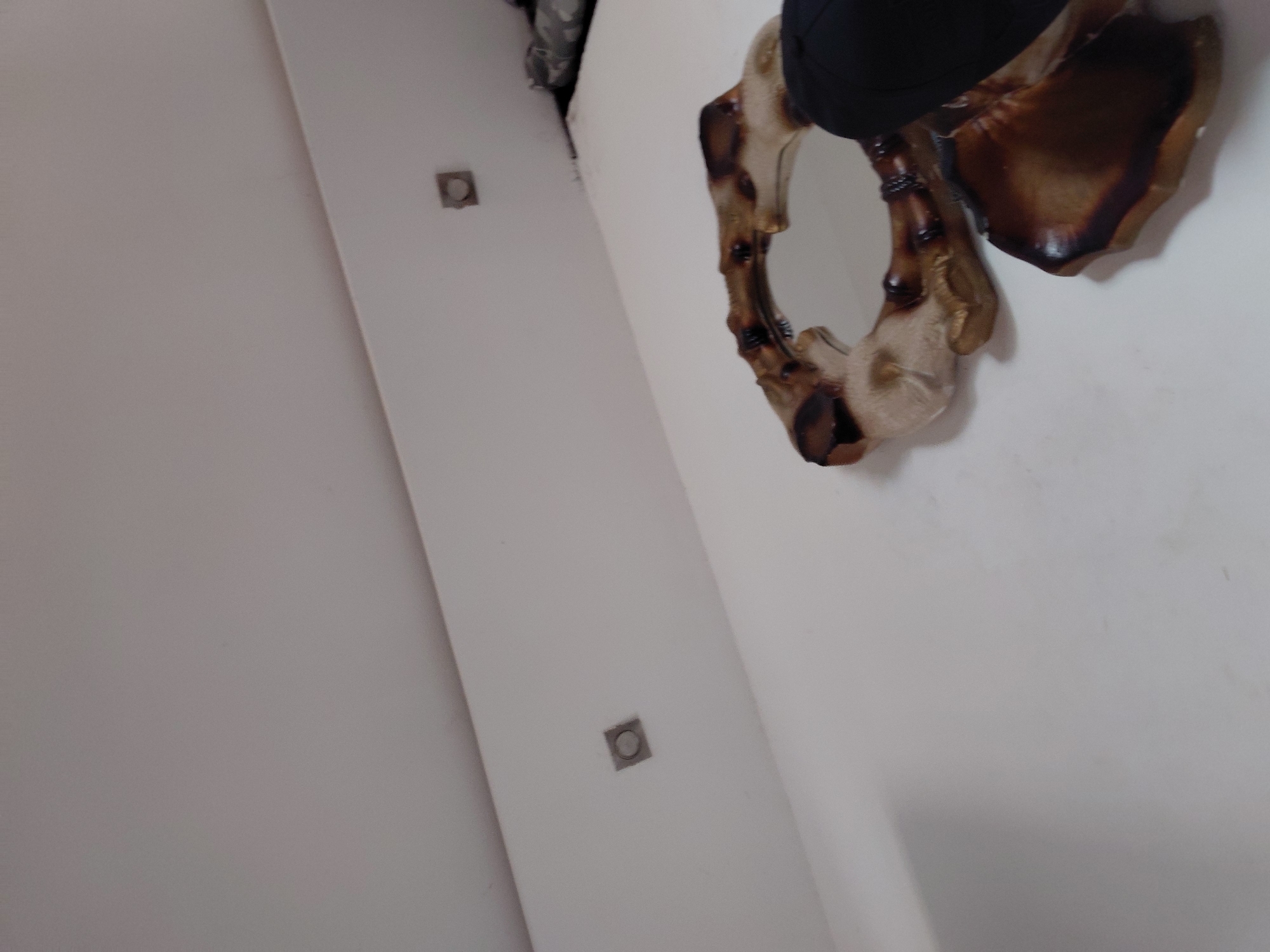 The image displays an indoor setting, likely a room with plain white walls, captured from a tilted, upward-looking perspective. The primary subject is a decorative mirror positioned in the upper right section of the frame. The mirror features a distinctive, organic-shaped frame that resembles carved wood, bone, or resin, exhibiting a mix of light, creamy beige tones and darker, rich brown, almost burnt-looking areas. The surface of the frame has a somewhat glossy finish, highlighting its natural textures and irregularities. The small, circular mirror itself reflects a dark, indistinct object, possibly a piece of clothing or part of another decorative item, rather than the room's interior.
On the white wall, two small, square fixtures are visible, positioned diagonally. These appear to be electrical outlets or utility access points, each featuring a circular indentation or component in its center. The wall surface is smooth and uniformly white. In the far upper left corner, out-of-focus dark shapes are minimally present, suggesting other structural elements of the room or objects out of view. The scene is static, with no discernible activity or people present. The lighting is indirect, typical of an indoor environment. There is no visible text in the image. This interior scene is located in Santa Ana, Venezuela.