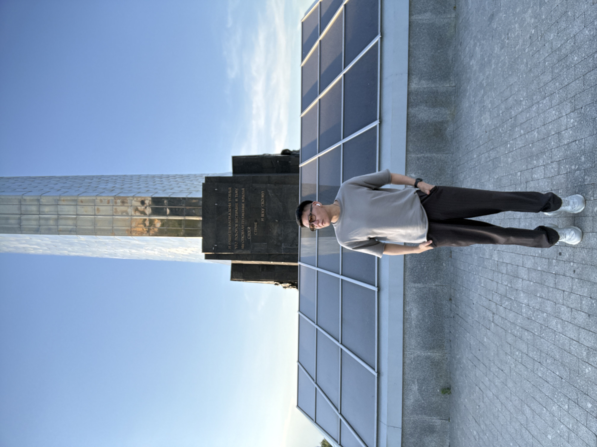 A young Asian man stands outdoors in front of a monument under a clear blue sky. He is wearing a grey t-shirt, dark striped pants, and white sneakers. He has on glasses and earbuds. The monument is tall and metallic, with a darker stone base. The man is positioned centrally in the frame, facing the viewer with his hands in his pockets. The ground is paved with grey interlocking bricks. The scene appears to be in Moscow, Russia, as the monument is the Conquerors of Space monument.