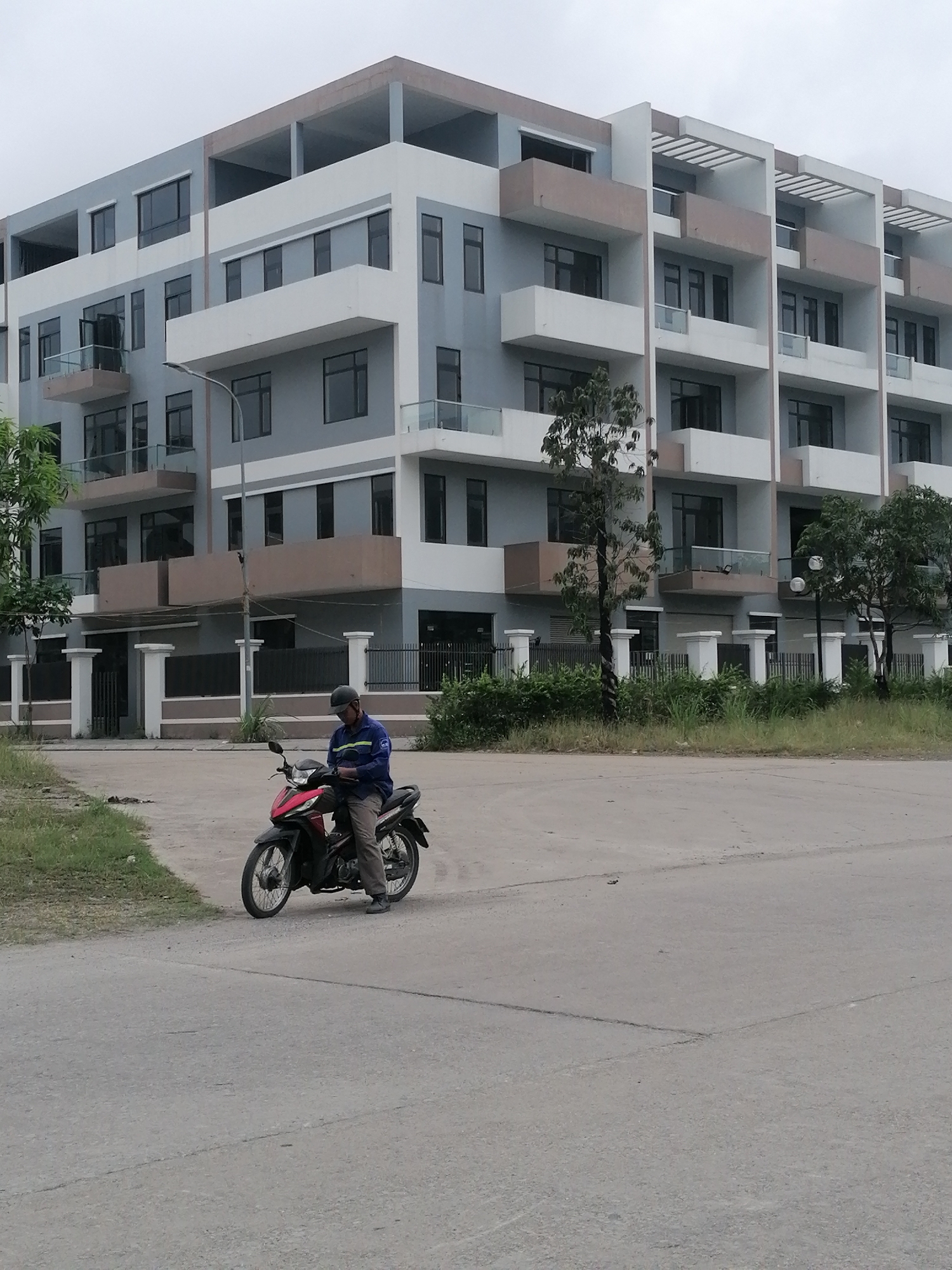 An outdoor, eye-level shot captures a modern residential or commercial area in Ha Tu Ward, Vietnam, on an overcast day. In the foreground, a man sits on a red and black motorbike on a concrete road. He wears a blue long-sleeved shirt, dark pants, and a dark helmet, appearing to be looking down at something near his hands. To the left of the road, there are patches of dry grass.
The background features a large, multi-story building with a contemporary design, characterized by a mix of light gray, white, and light brown architectural elements. It has numerous dark-framed windows and balconies with glass railings. The top floor appears to be an open-air structure. A white-pillared fence with dark metal railings runs along the base of the building, separating it from the road. Scattered green bushes and small trees are visible along the fence line and adjacent to the building. The sky is uniformly gray, indicating an overcast day. No discernible text is visible in the image.