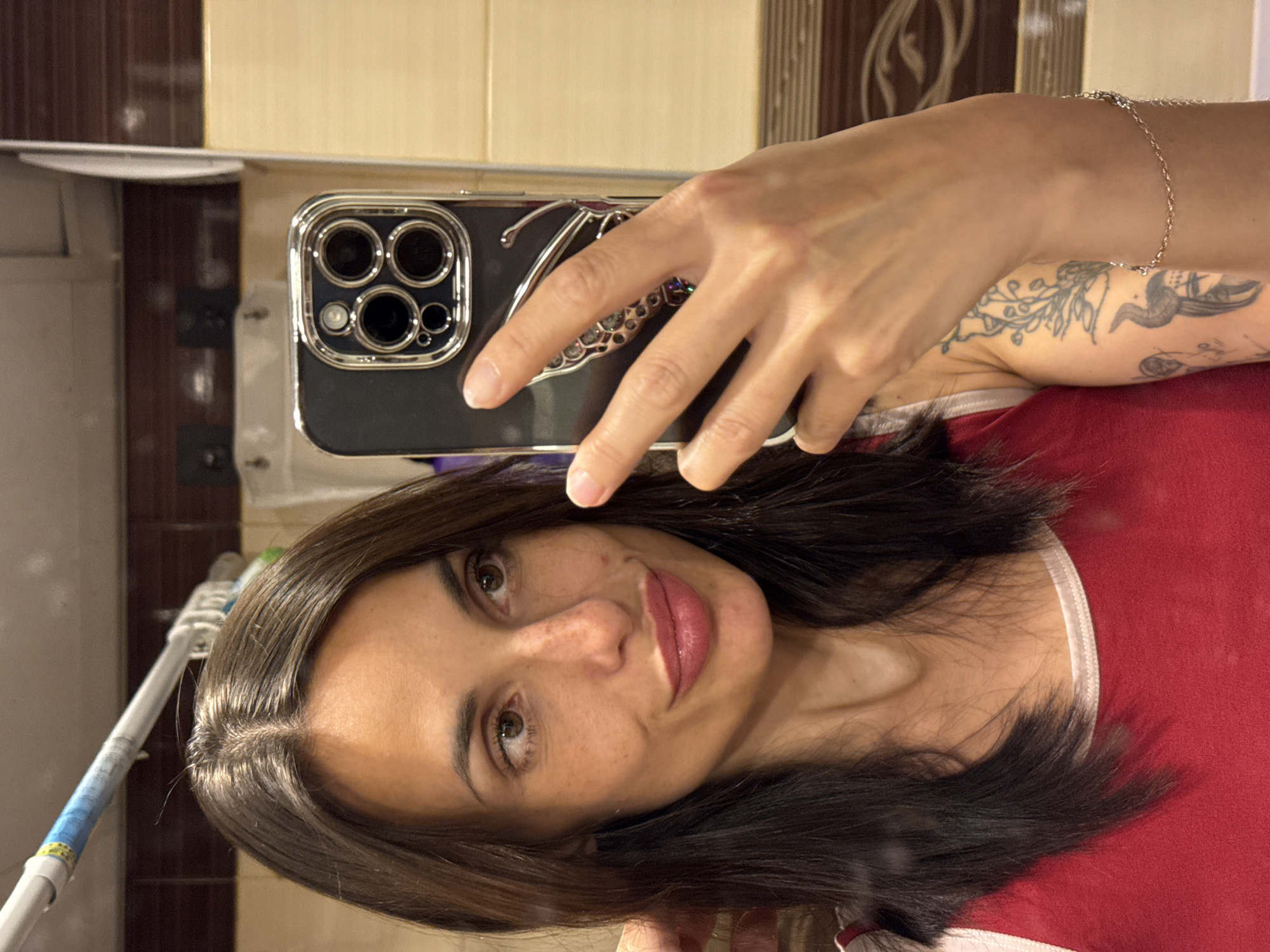 A woman with dark hair and freckles takes a mirror selfie in a bathroom. She is wearing a red tank top with white trim. Her hair is dark brown and falls to her shoulders. She has brown eyes and is wearing lip gloss. In her right hand, she holds a smartphone with a black case and a silver butterfly charm. A silver bracelet is visible on her right wrist. Behind her, the bathroom walls are tiled in shades of brown and beige. A white shower rod with blue tape is visible above her head. The image appears to have been taken in the city of Pleven, Bulgaria, as indicated by the original prompt.