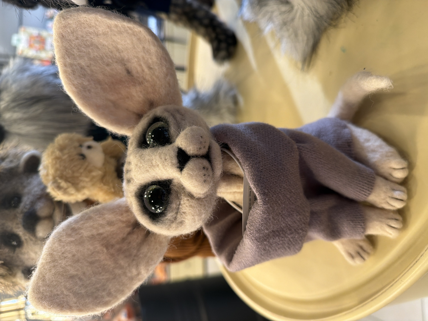 An overhead, slightly tilted close-up shot captures a charming, handcrafted felted animal toy resting on a light yellow surface. The main subject is a light grey or beige creature, possibly a fennec fox or a rabbit due to its notably large, elongated ears. It features prominent, glossy black eyes that reflect light, giving it a bright, engaging gaze, and a small, pointed snout with dark nostrils. The toy is wearing a snugly fitted, long-sleeved knitted sweater in a muted purple or mauve color, which covers its body and part of its limbs. Its felted paws, complete with visible toes, extend from the sweater's cuffs and are neatly placed in front. A small, lighter-colored felted tail is visible behind the sweater.
In the blurred background, other similar animal toys are visible, suggesting a retail or display setting like a craft store, boutique, or market stall. To the left, a smaller, lighter brown fluffy toy is partially seen, and below it, part of a grey felted head, possibly a raccoon or similar animal, is out of focus. The overall scene is brightly lit, likely by indoor artificial lighting, making the time of day and external weather indeterminate. No discernible text is visible on the toy, its sweater, or any tags. The craftsmanship of the felted toy is evident, creating a cute and appealing display item from Saint Petersburg, Russia.