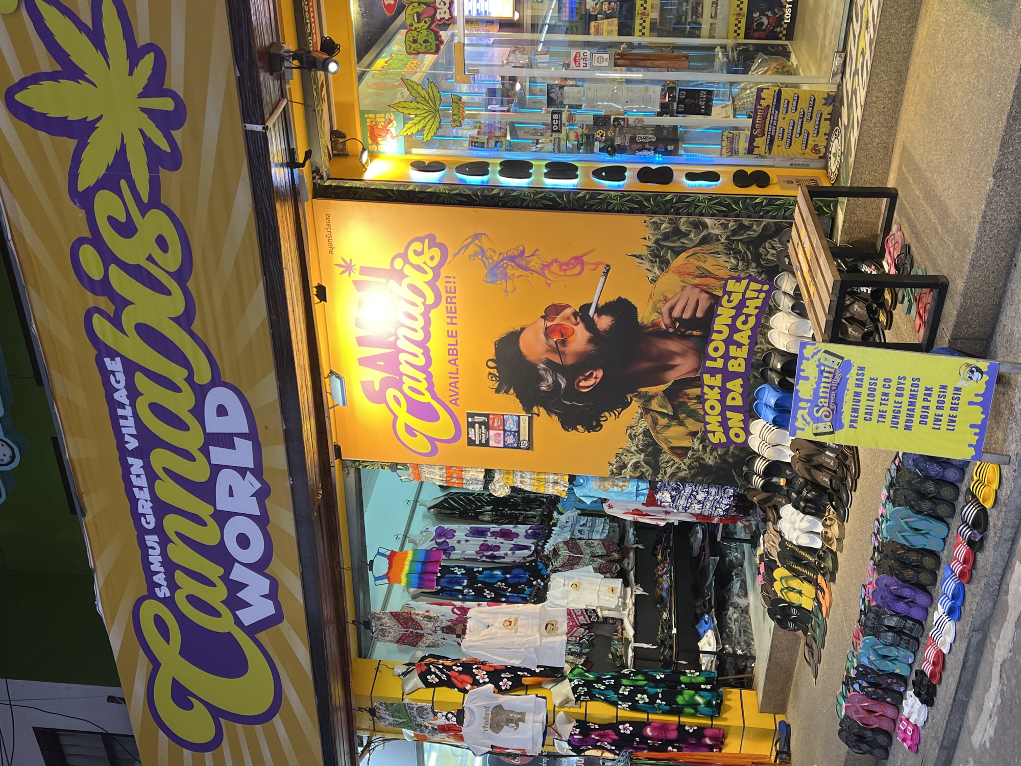 This image captures the exterior of a cannabis shop in Baan Bo Phut, Thailand. A large yellow banner dominates the scene, featuring the words "Cannabis World" in bold purple and yellow lettering. Below this, a striking poster depicts a person with sunglasses and a cigarette, with the text "SMOKE LOUNGE ON DA BEACH!!" above it. The shop's interior is visible through a glass window, displaying shelves stocked with various products, many of which appear to be cannabis-related items, with neon blue lighting illuminating the display.
In front of the shop, a selection of clothing items, including t-shirts and patterned fabrics, are displayed on racks. A wooden bench is placed on the sidewalk, and a variety of flip-flops are neatly arranged on the ground in front of the store, suggesting a casual, beachside atmosphere. The overall impression is that of a retail establishment catering to tourists, likely in a tropical or sub-tropical setting given the clothing styles and the "on da beach" slogan. The lighting suggests it might be evening or nighttime, with the shop's lights contributing to the ambiance. The visible text indicates the shop's name, "Samui Green Village," and lists various cannabis products like "Premium Hash," "Edibles," and "Live Rosin," along with prices and potential strain names. The presence of a Thai script further confirms the location.