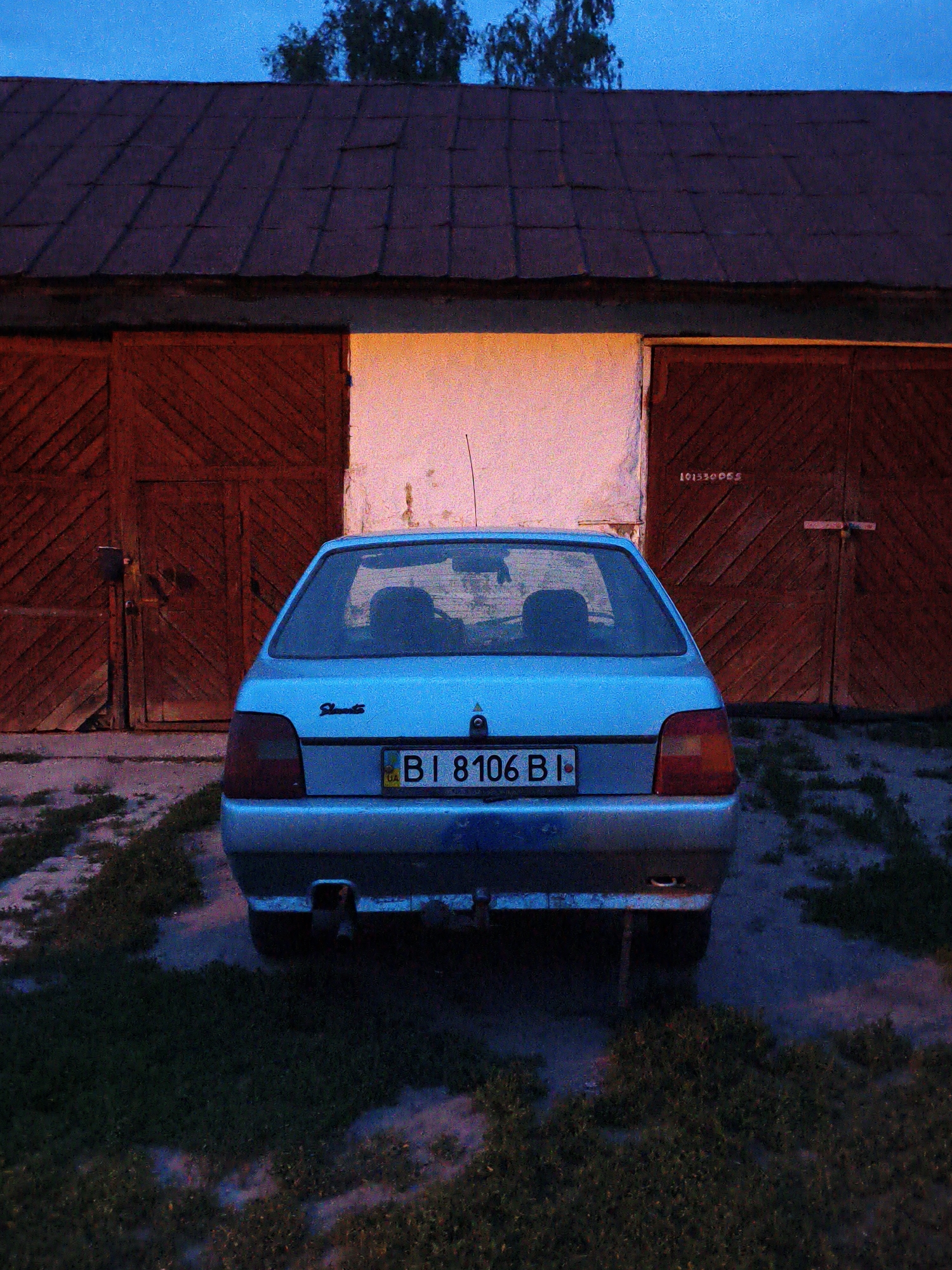 An outdoor, eye-level shot captures the rear view of a parked light-blue hatchback car against a rustic garage building at dusk or early evening in Ustyvytsia, Ukraine. The sky above is a deep blue, with the silhouette of distant tree foliage visible at the top.
The central subject is a silver-blue car, identified by a "Slavuta" badge on the left side of its trunk, parked on a patch of grass and dirt. Its rear window reflects a blue hue from the sky, and vague silhouettes of headrests are visible inside. The car bears a Ukrainian license plate that reads "BI 8106 BI," with the country code "UA" visible on the left. A small exhaust pipe is visible beneath the rear bumper.
Behind the car, a wooden garage building with a dark, possibly slate or metal, gabled roof dominates the background. The building features three sections: two dark red wooden double garage doors on either side, flanking a brightly lit central opening or section. The light emanating from this central opening is a warm, orange-yellow hue, strongly contrasting with the cool blues and dark reds of the scene. The wooden doors have diagonal plank patterns. On the right garage door, faint white text reads "10130088". The area directly in front of the building is a mix of concrete or packed earth and sparse green grass, which also extends to the foreground where the car is parked. The overall lighting suggests twilight, with shadows lengthening and artificial light beginning to stand out.