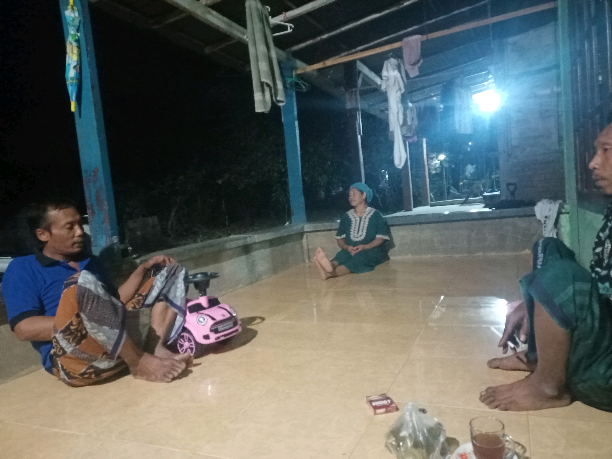 In Bubulan, Indonesia, a group of people are gathered outdoors at night. A man wearing a blue shirt and patterned pants is seated on the floor, next to a small pink toy car. Another person, possibly a woman, is seated further back, wearing a green headscarf and a matching green garment. To the far right, another individual is partially visible, wearing a dark garment and green pants. The scene appears to be on a tiled porch or patio area, with a dark background suggesting trees or foliage. Clothes are hanging on a clothesline above. A light source, possibly a lamp, illuminates the area from the right. A pack of cigarettes is on the floor, along with a glass of liquid.