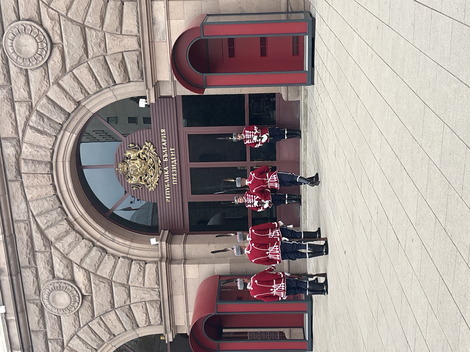 A ceremonial guard stands in formation outside a stately stone building in Sofia, Bulgaria. Five soldiers, clad in distinctive red jackets with white braiding, dark trousers, black boots, and hats adorned with white feathers, are positioned across the paved entrance area. Each soldier holds a rifle with a fixed bayonet.
The building's facade is constructed from light-colored, roughly cut stone blocks, featuring an imposing arched main entrance framed in dark wood with reflective glass. Above the entrance, a prominent gilded national coat of arms is displayed. To the right of the coat of arms, Cyrillic text is clearly visible, spelling out "РЕПУБЛИКА БЪЛГАРИЯ ПРЕЗИДЕНТ" (Republic of Bulgaria President).
Flanking the central entrance are two modern, curved guard boxes or shelters, predominantly red with dark trim. One soldier is visible standing inside the rightmost guard box. The ground is paved with large, light-colored flagstones. Architectural details on the building include circular carved medallions embedded in the stone facade. The lighting suggests a bright day, possibly overcast, as shadows are diffused. The scene captures a moment of official duty at the Bulgarian Presidency.